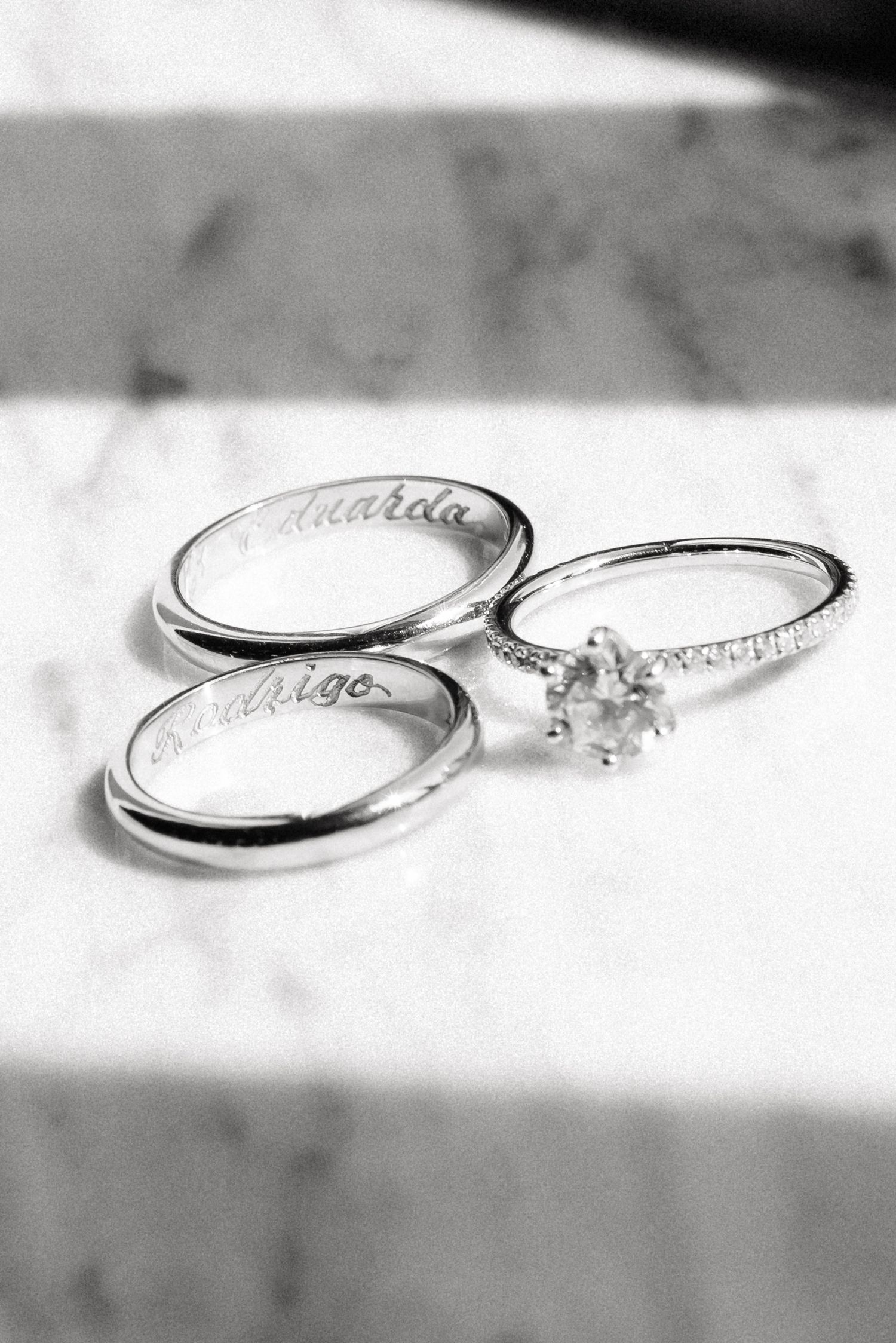 Black and white photo of three silver wedding rings engraved with names Eduarda and Rodrigo on marble surface.