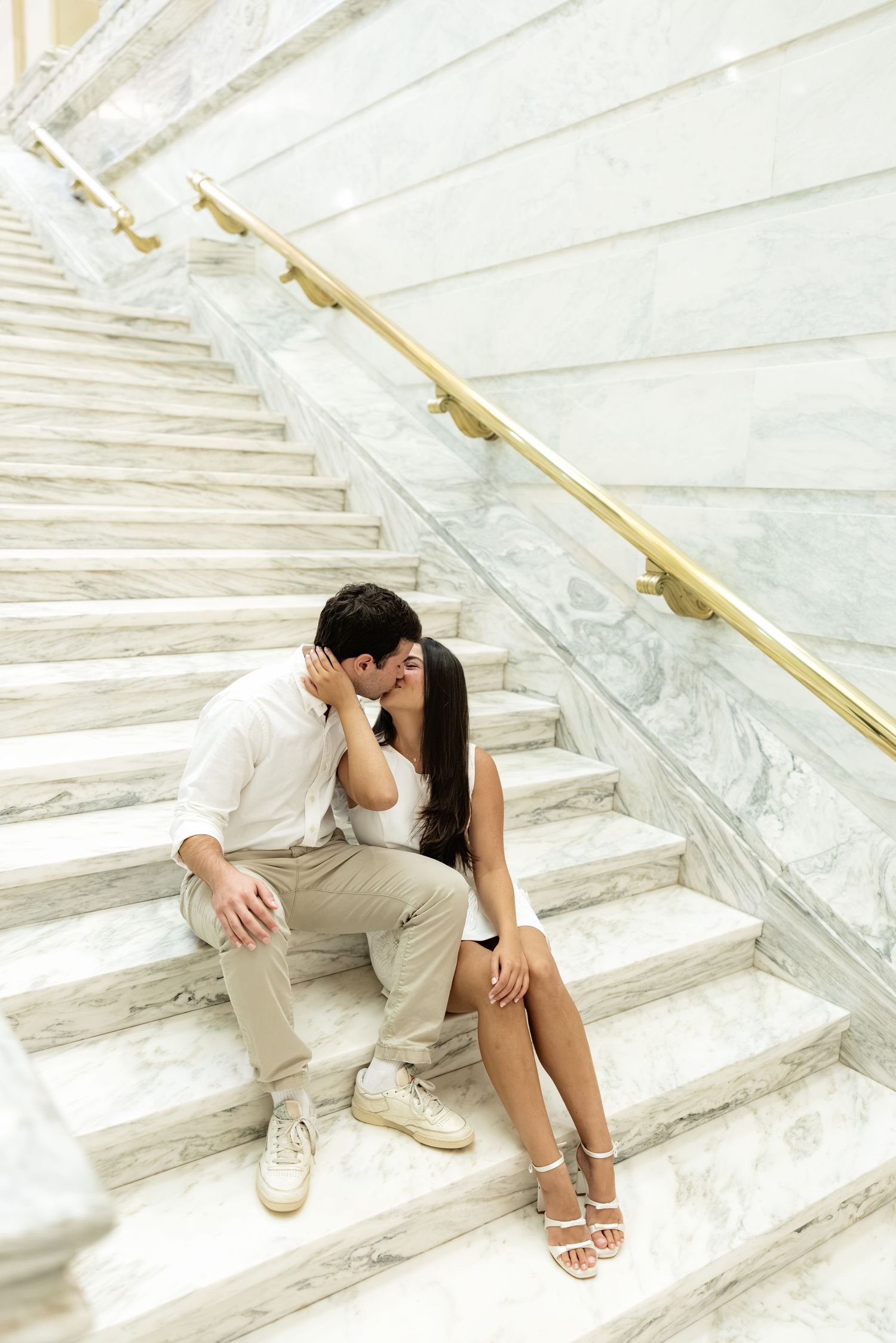 Romantic couple sharing a kiss while sitting on elegant marble stairs with gold railings in a luxurious building.