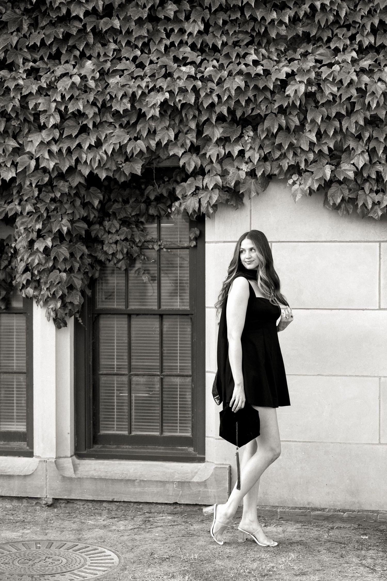 Black and white portrait of a woman in a black dress standing by an ivy-covered building.