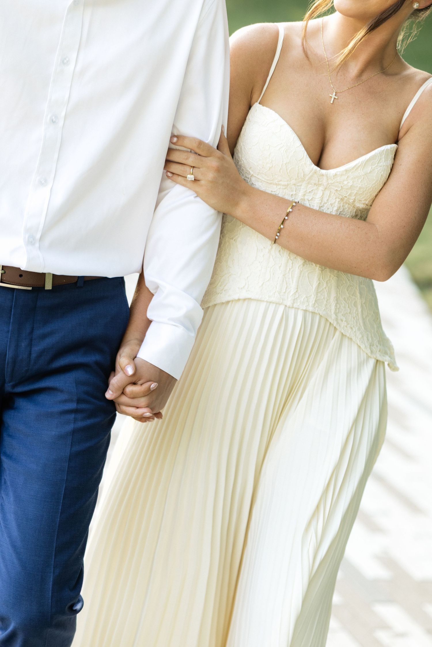 Couple holding hands, woman in cream pleated dress with cross necklace, man in white shirt and navy pants.