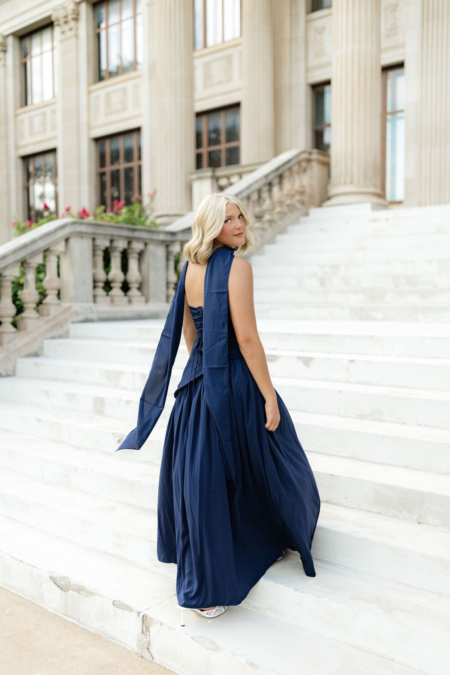 Blonde woman in elegant navy blue maxi dress with flowing scarf, posing on grand marble staircase.
