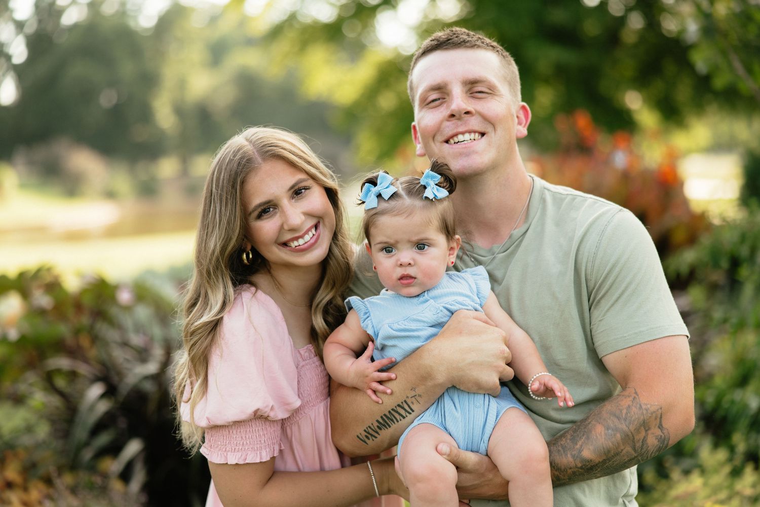 Happy young family with mom, dad, and baby girl posing outdoors in a lush green park setting.