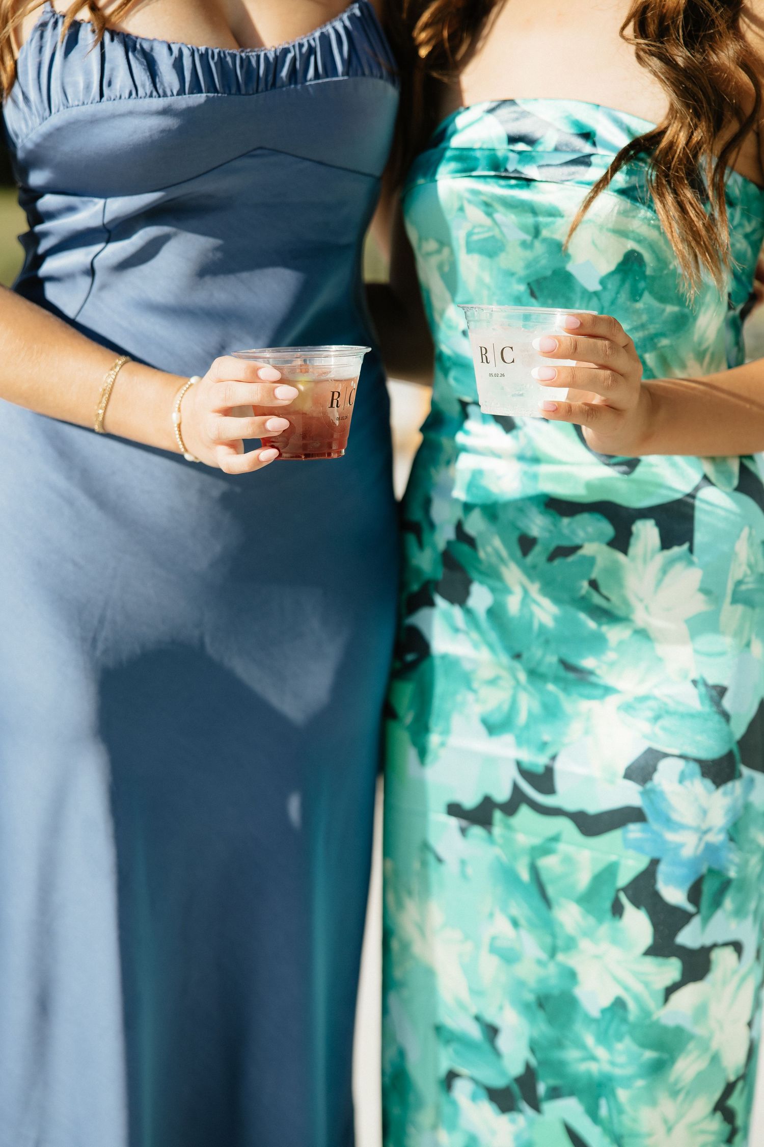 Two women in blue and floral green dresses holding monogrammed cocktail cups at a wedding celebration.