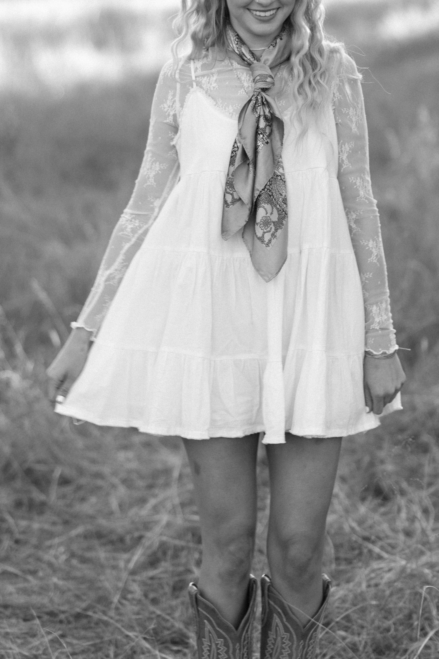 Young woman in white lace dress with neck scarf and cowboy boots standing in a field, black and white.