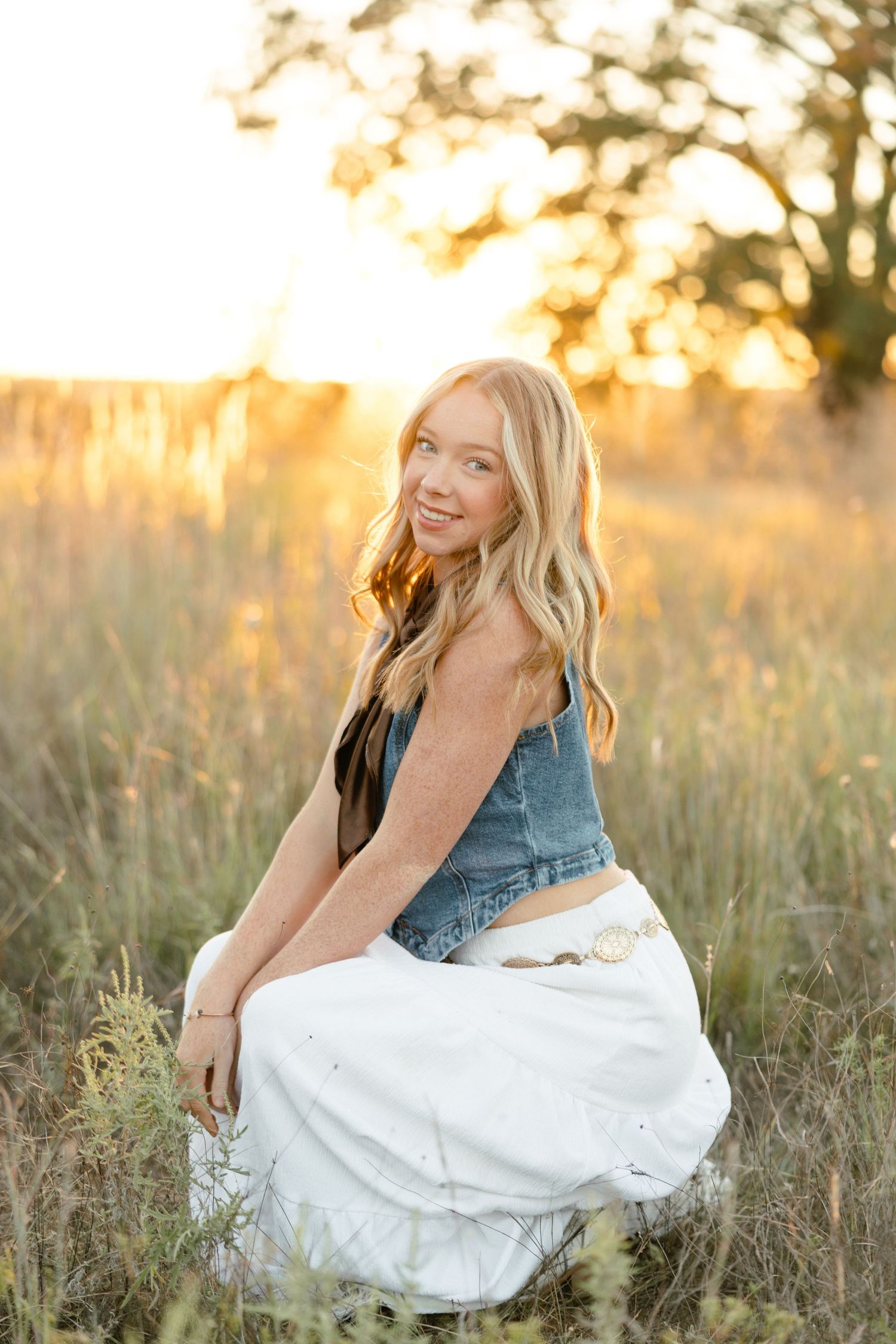 Smiling blonde teen girl in denim vest and white skirt sitting in a golden sunlit field at sunset.