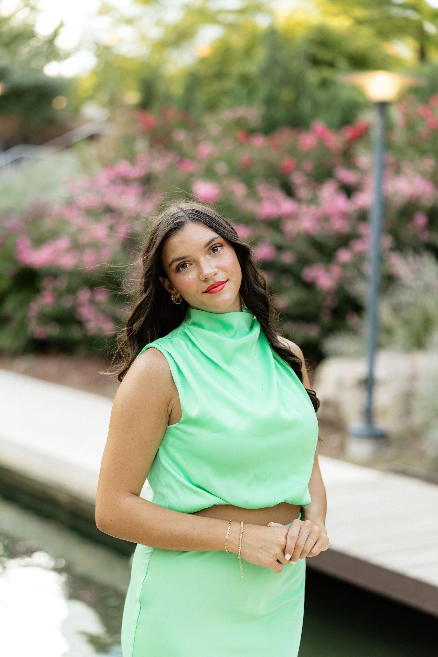 Young woman in mint green sleeveless turtleneck dress poses outdoors near water with pink flowering bushes behind her.