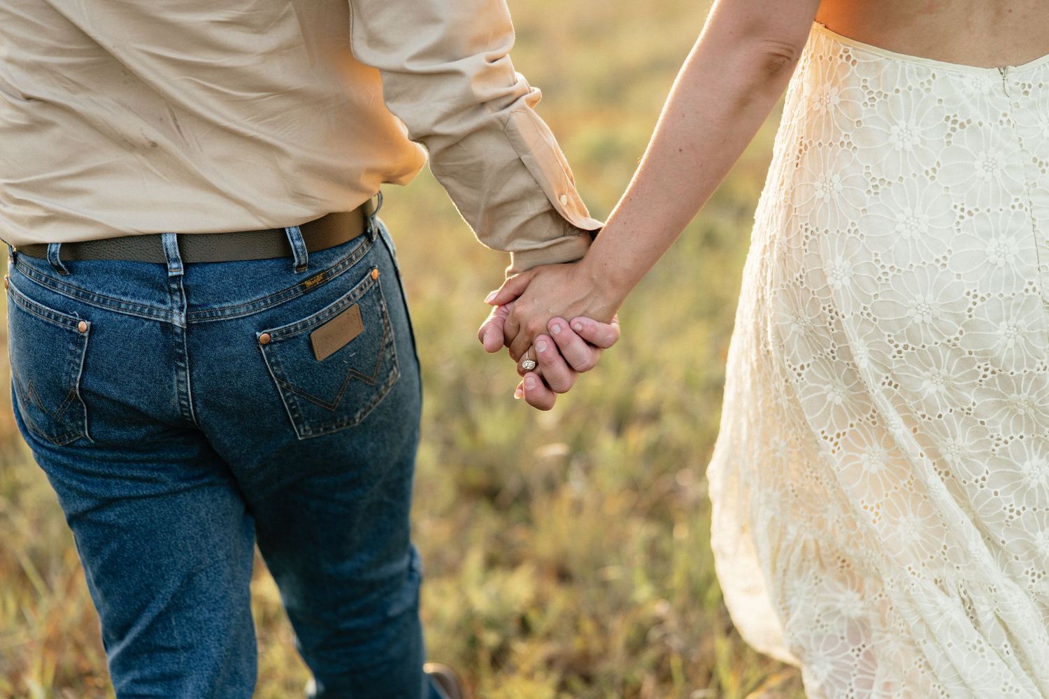 Close-up of an engaged couple holding hands in a field, wearing jeans and a lace dress.
