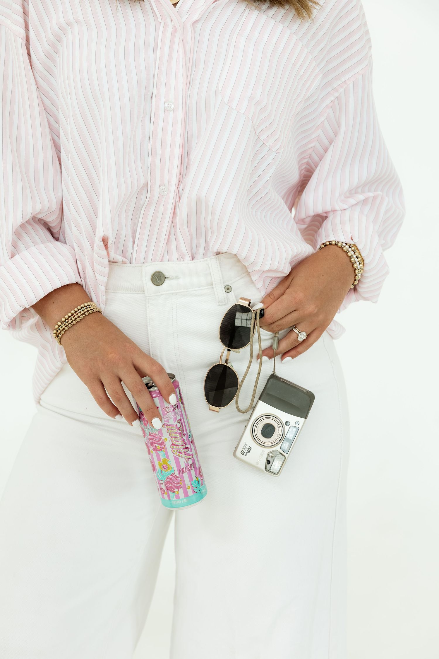Woman in white pants and pink striped shirt holding a colorful tumbler, sunglasses, and a retro camera.