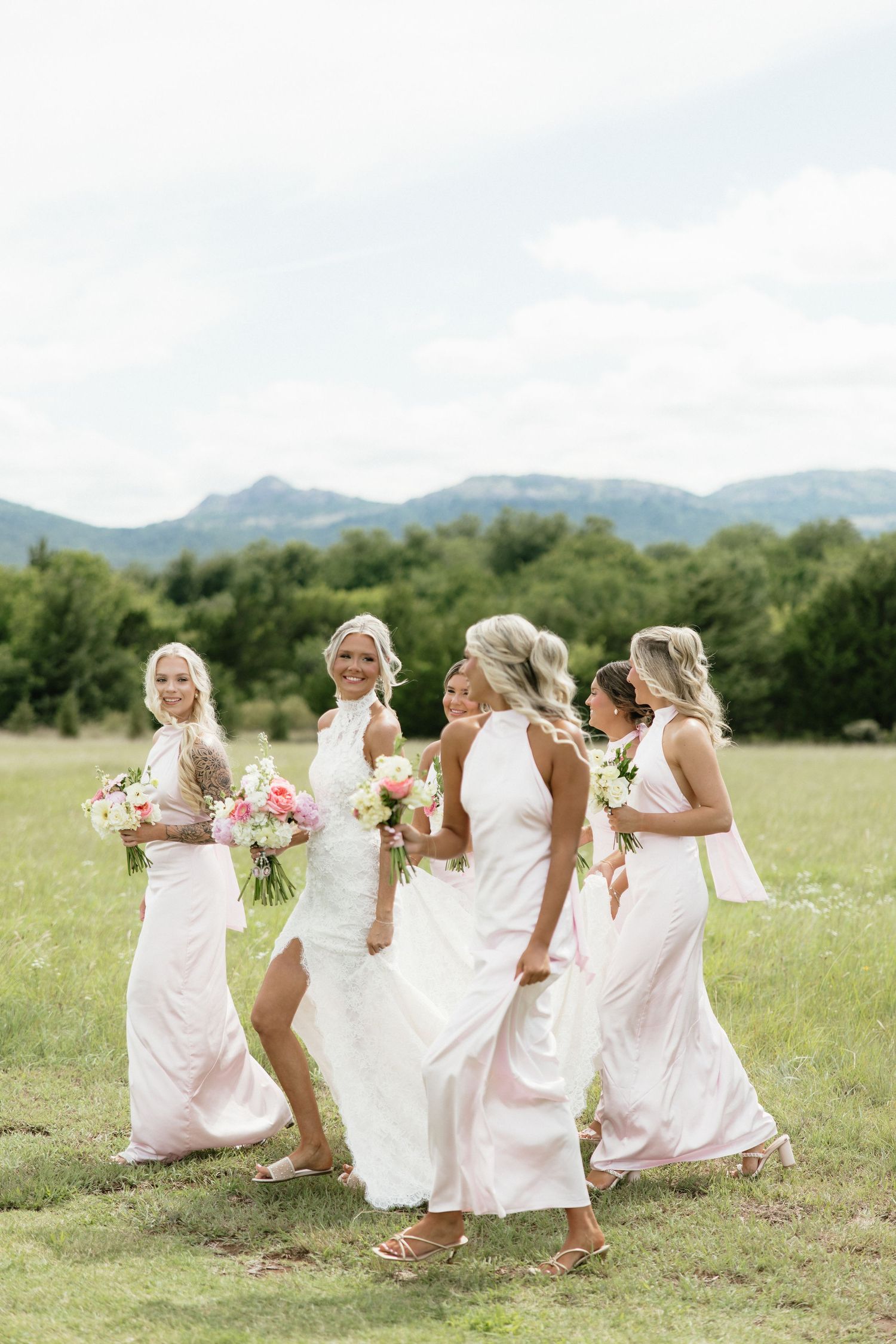 Bride walks with four bridesmaids in blush gowns holding floral bouquets in a green field with mountains behind.