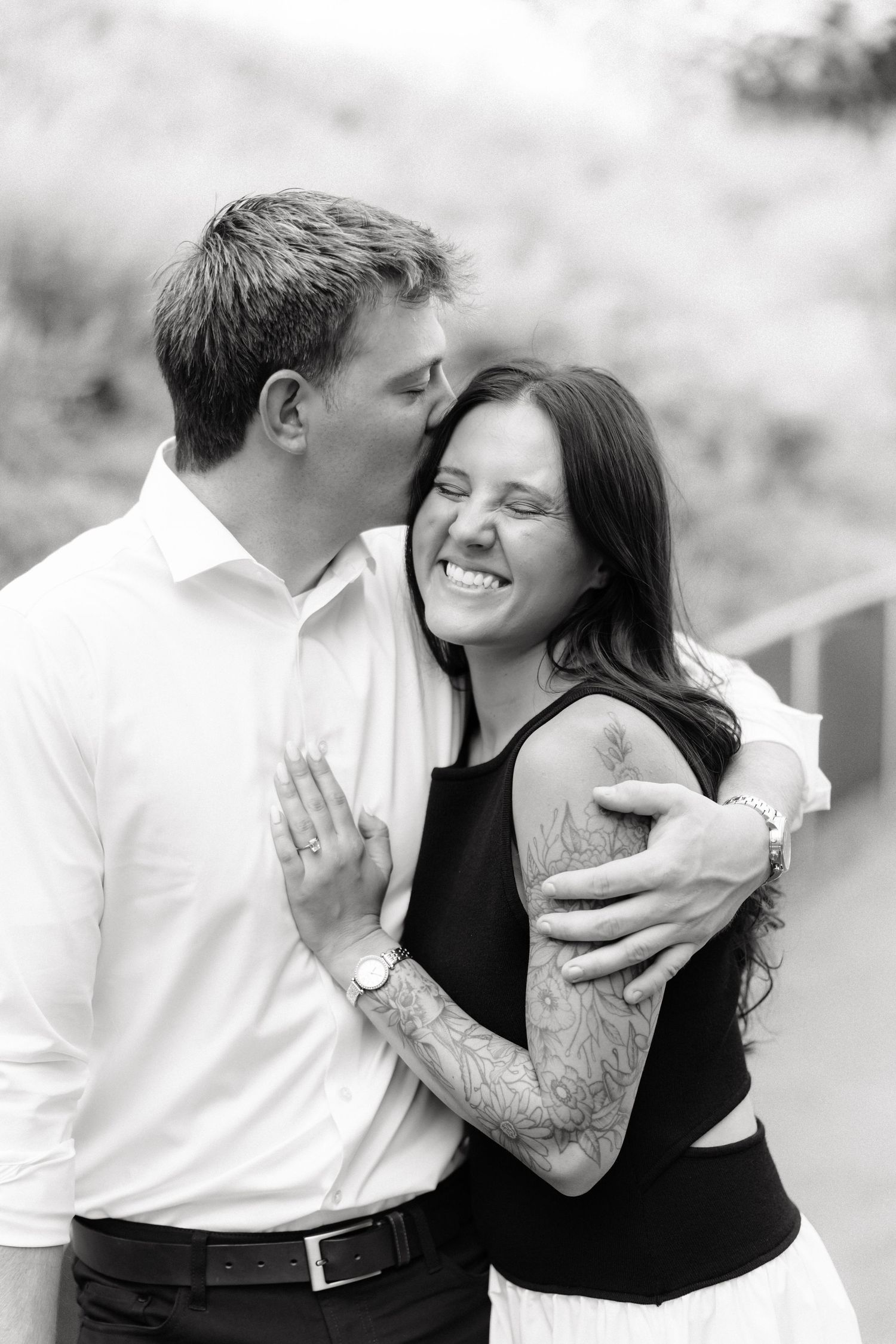 Black and white photo of a smiling couple embracing outdoors, man kissing woman's cheek, woman with tattoo on arm.