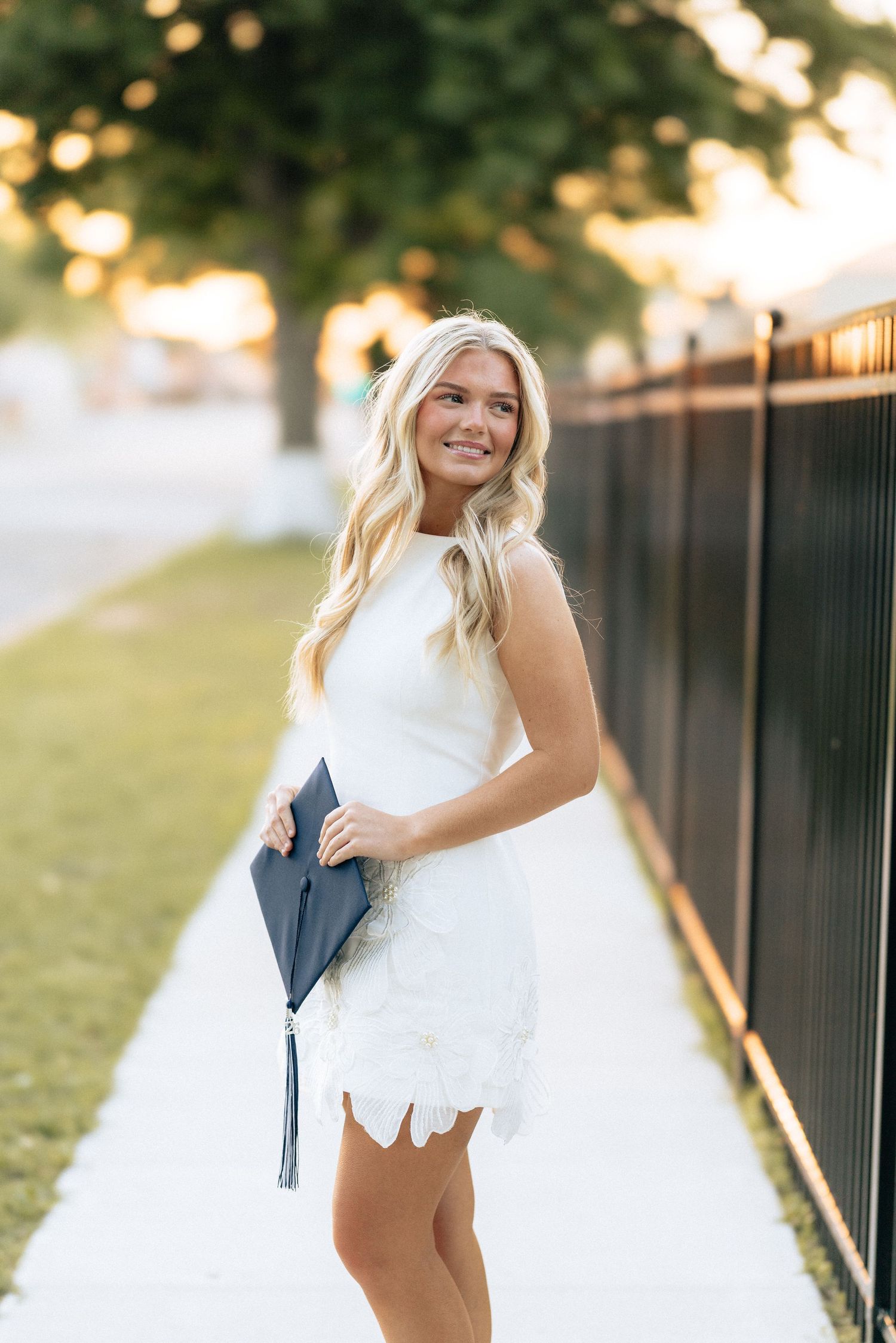 Blonde woman in white lace dress holding a graduation cap while smiling on a sunny sidewalk.