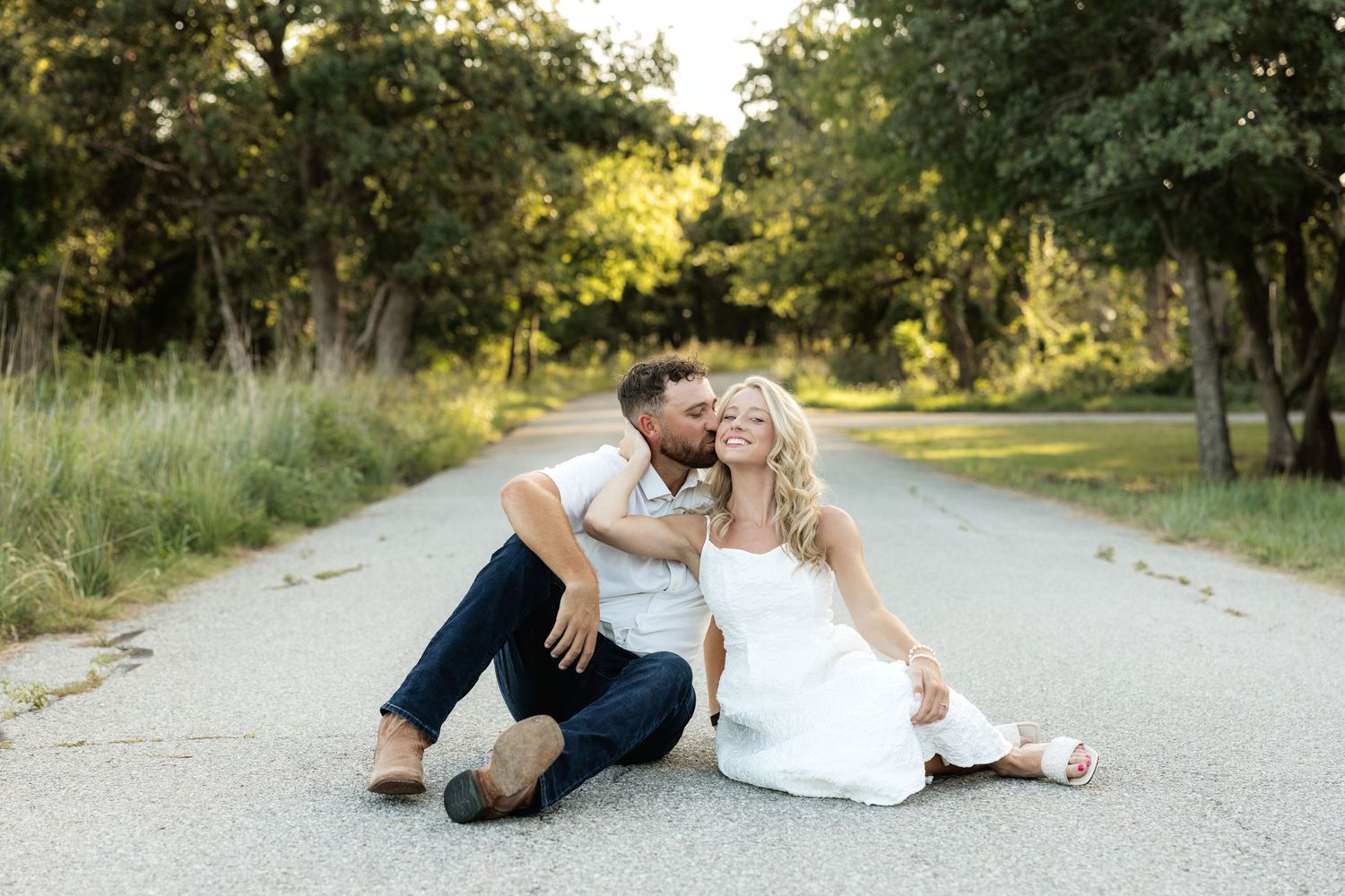 Couple sitting together on a tree-lined road, man kissing woman's cheek, both dressed in white and navy.