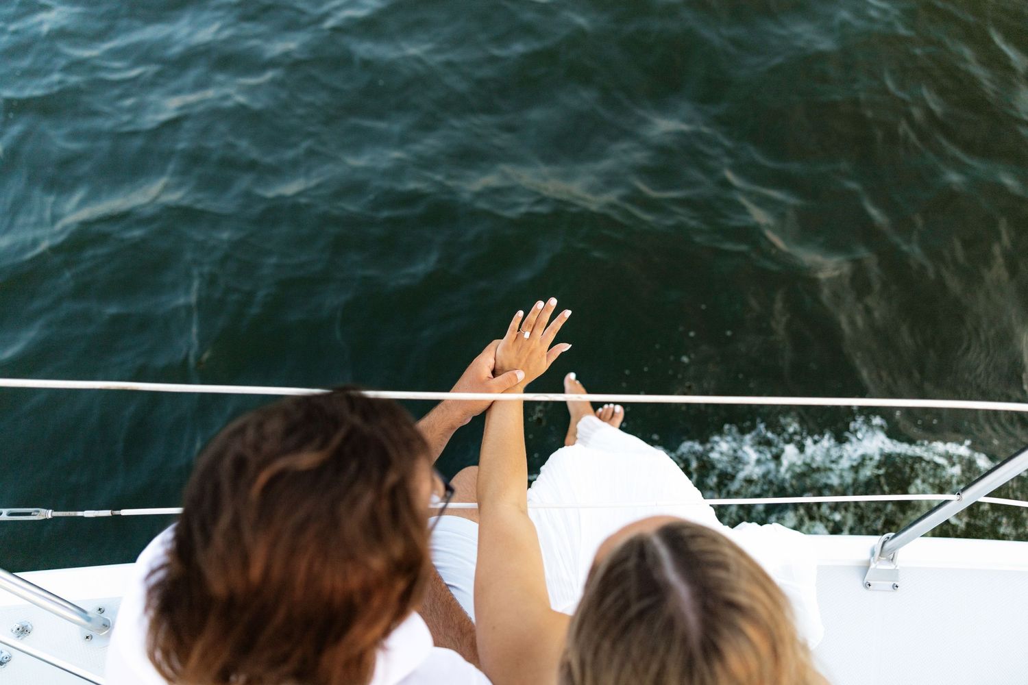 Two women on a yacht bow wave hands over ocean water on a sunny day.