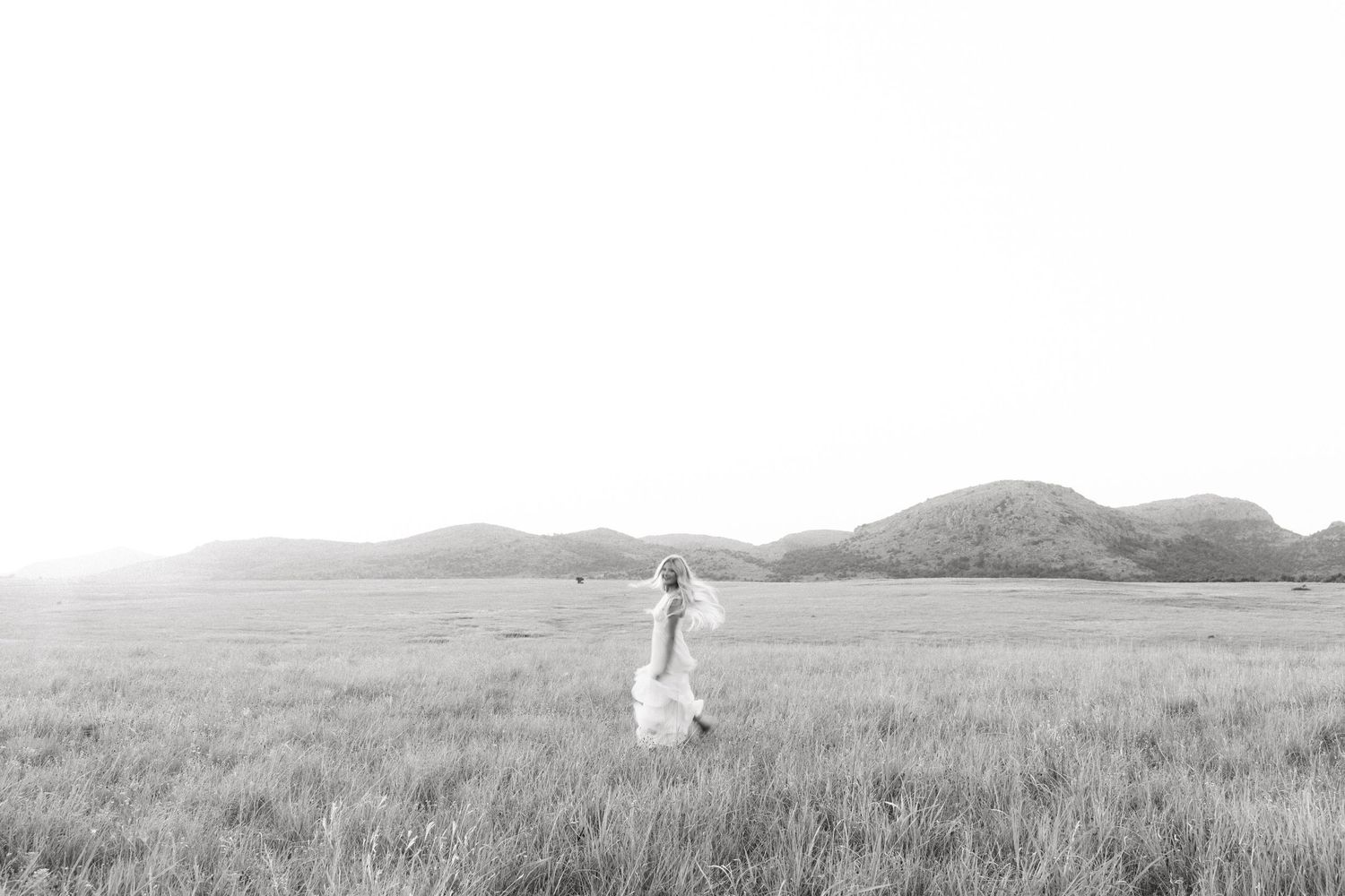 Black and white photo of a bride in a flowing white dress standing in an open field with mountains in the background.