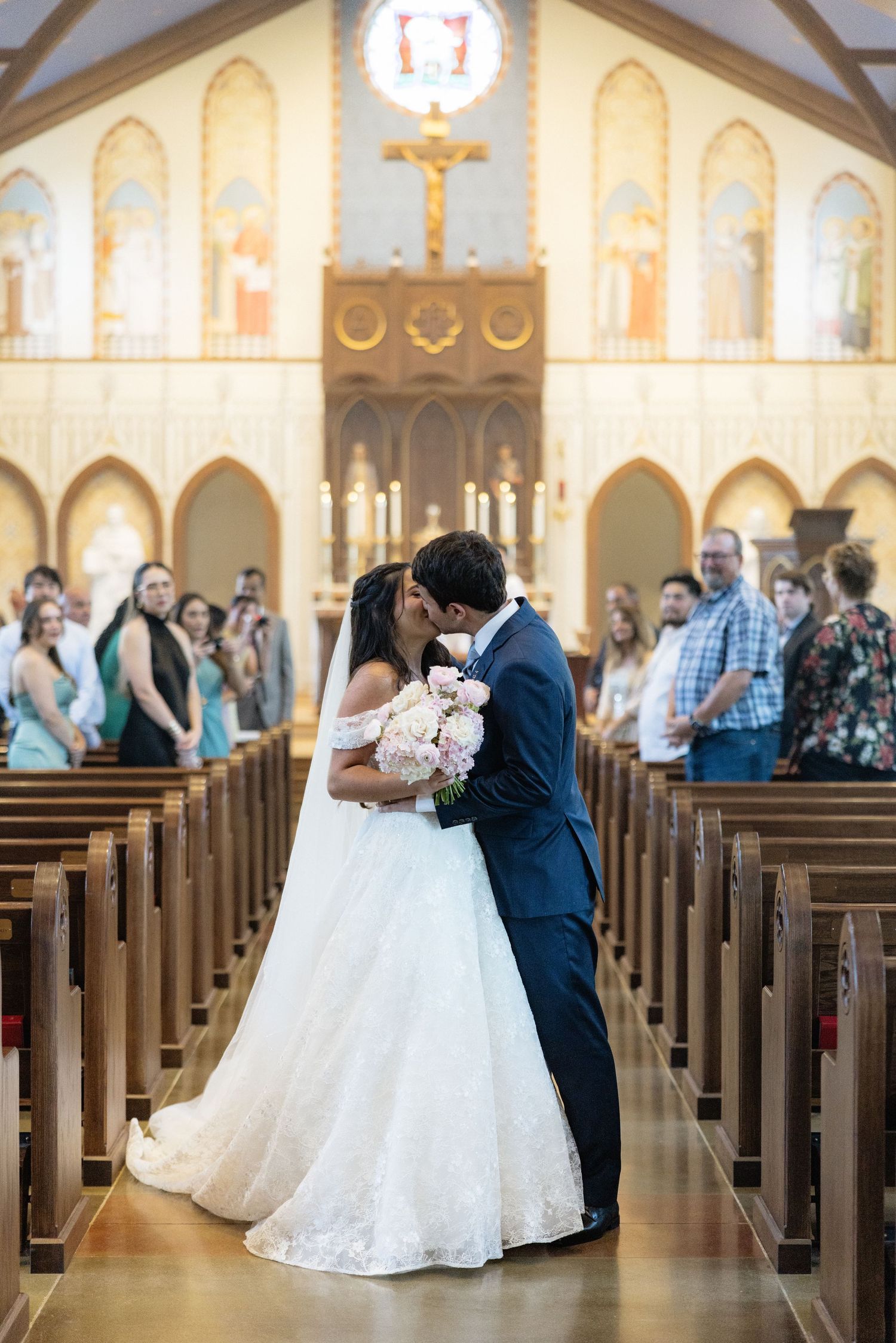Bride and groom share first kiss in church aisle, surrounded by guests, with altar and crucifix in background.