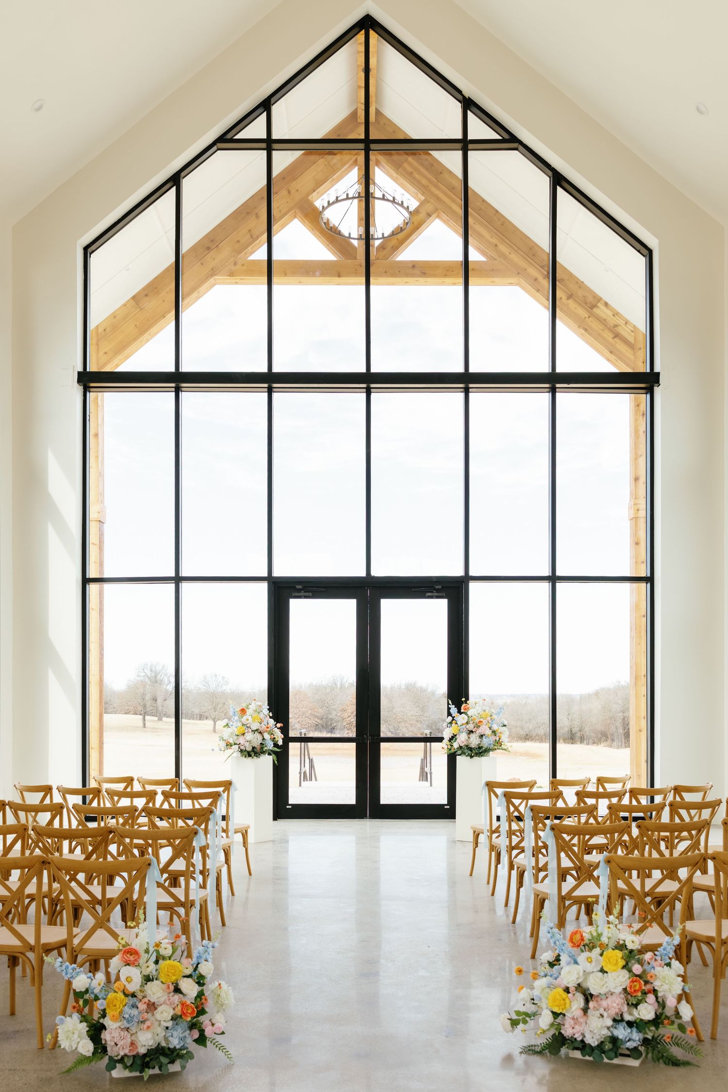 Bright modern wedding chapel with floor-to-ceiling glass windows, wooden beams, and colorful floral aisle arrangements.
