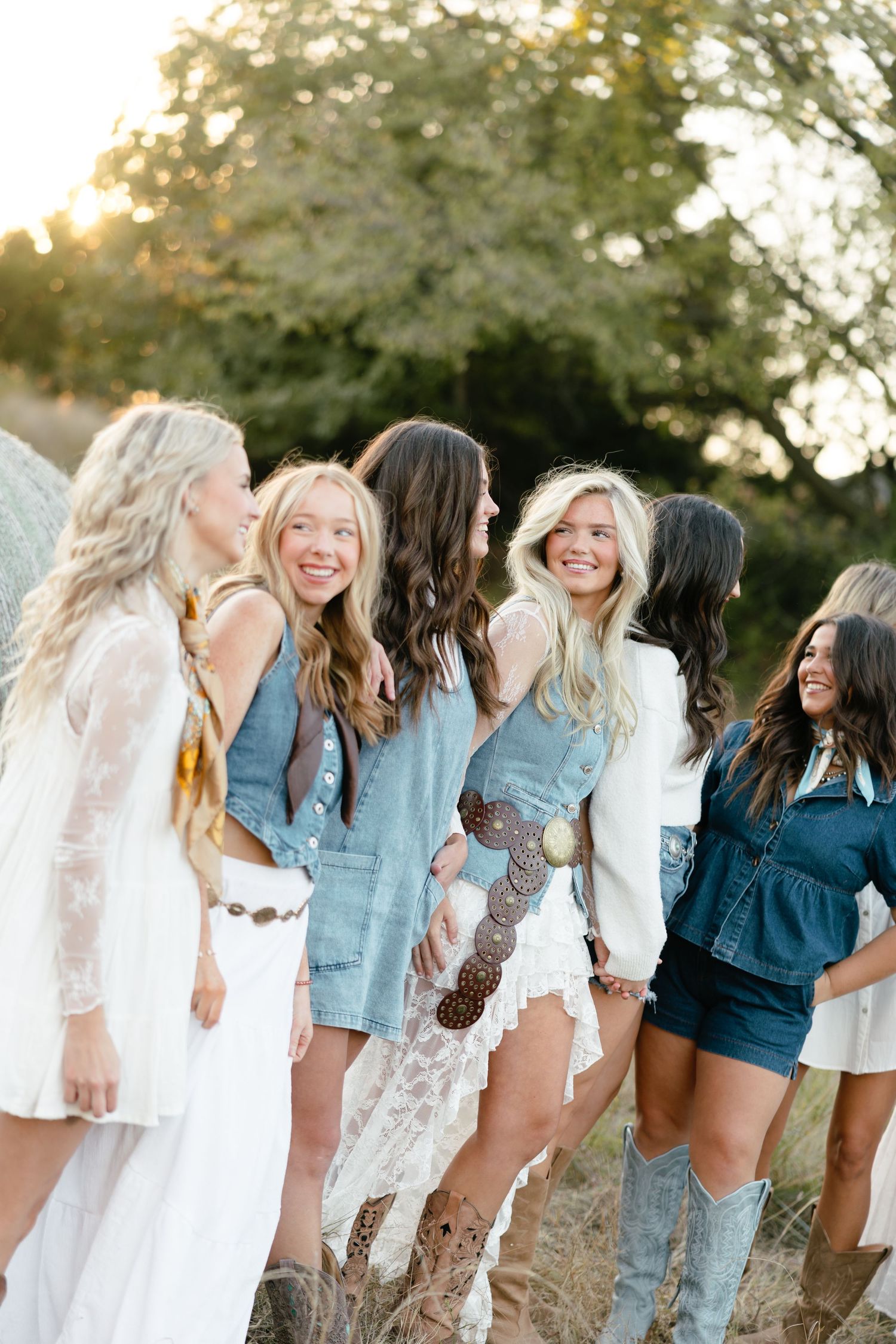 Group of smiling young women in boho western style outfits laughing together in a sunny field.