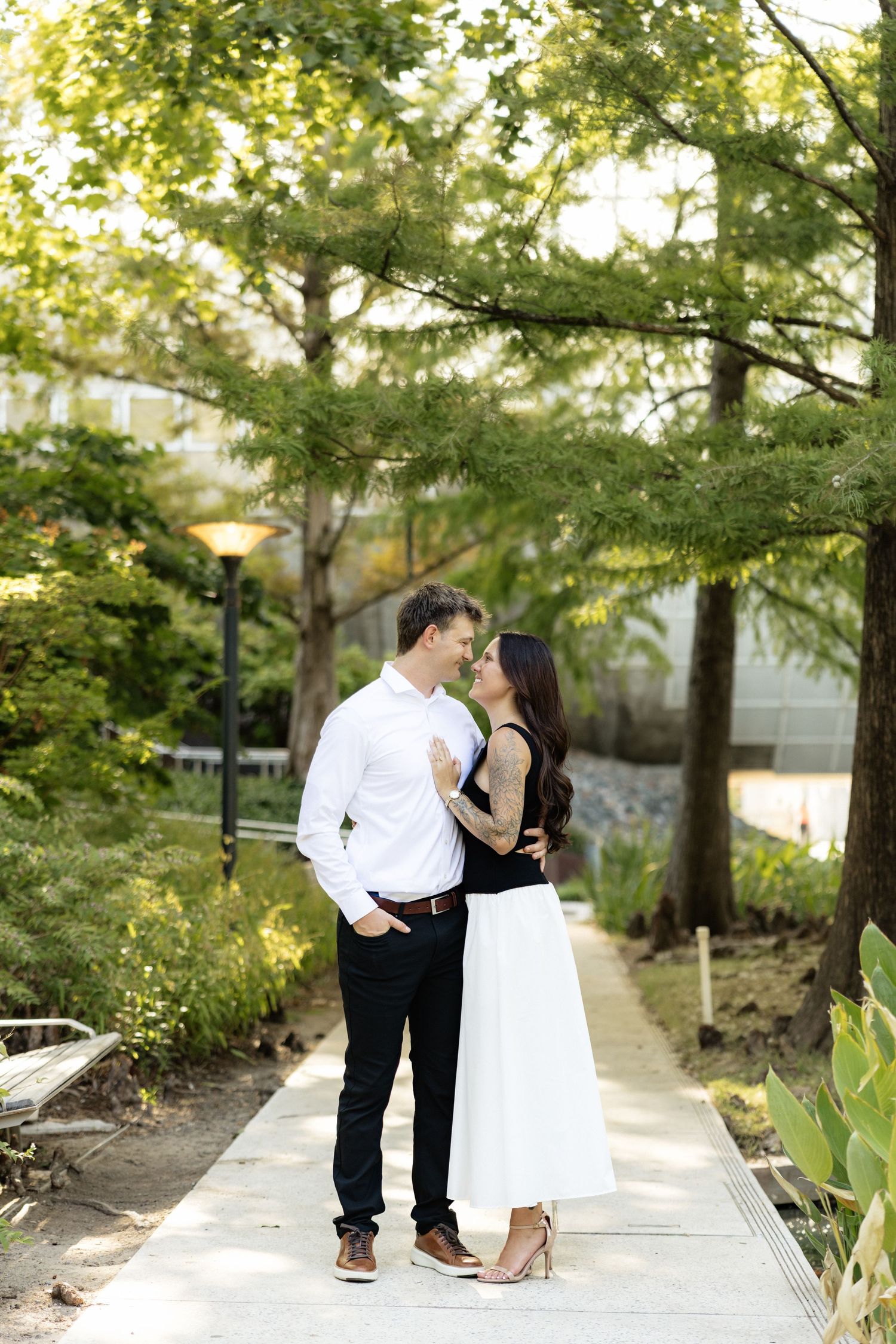 A couple shares a romantic kiss on a garden path surrounded by lush green trees in an outdoor park setting.
