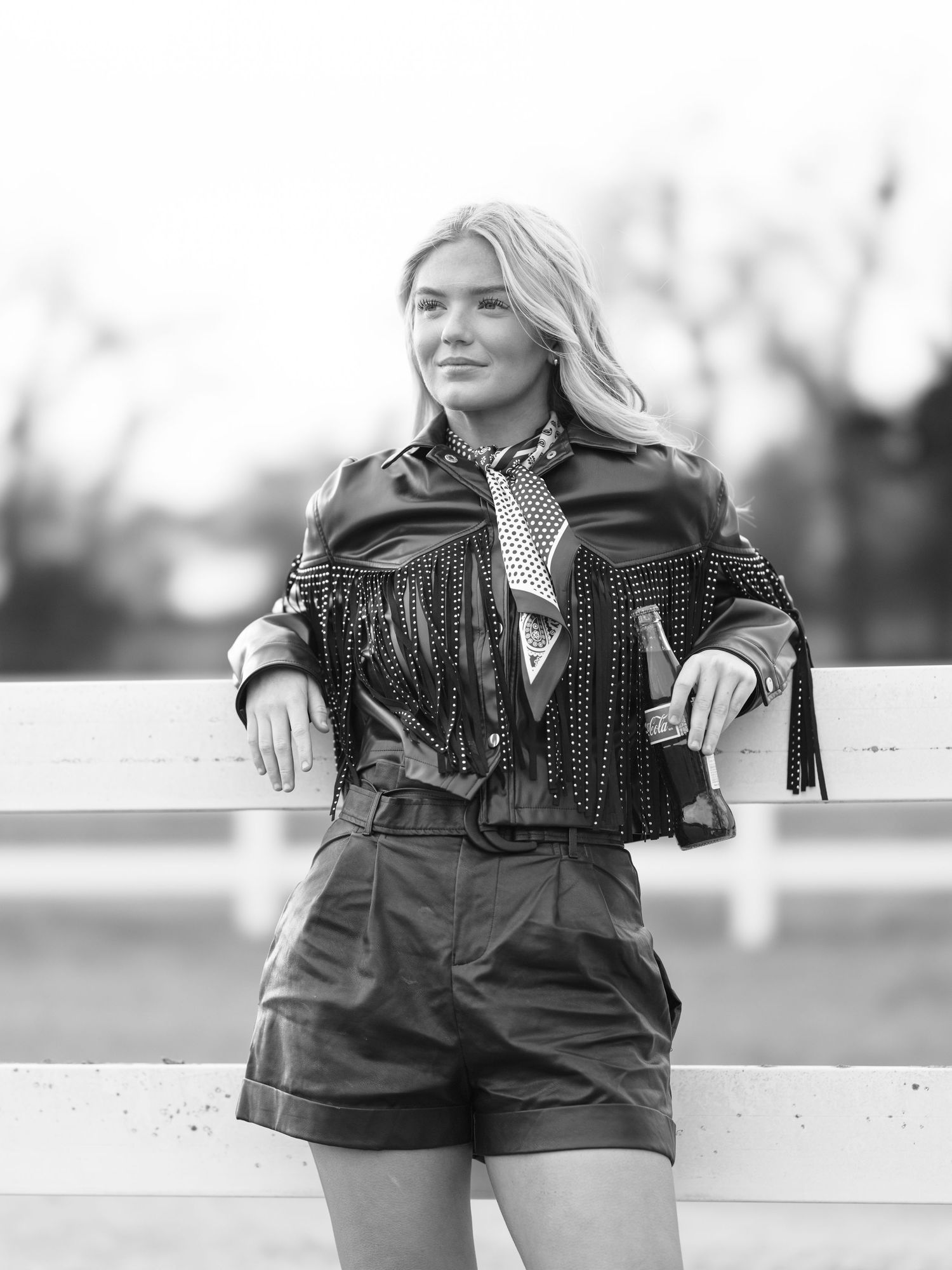 Black and white portrait of blonde woman in studded western jacket leaning on white fence.