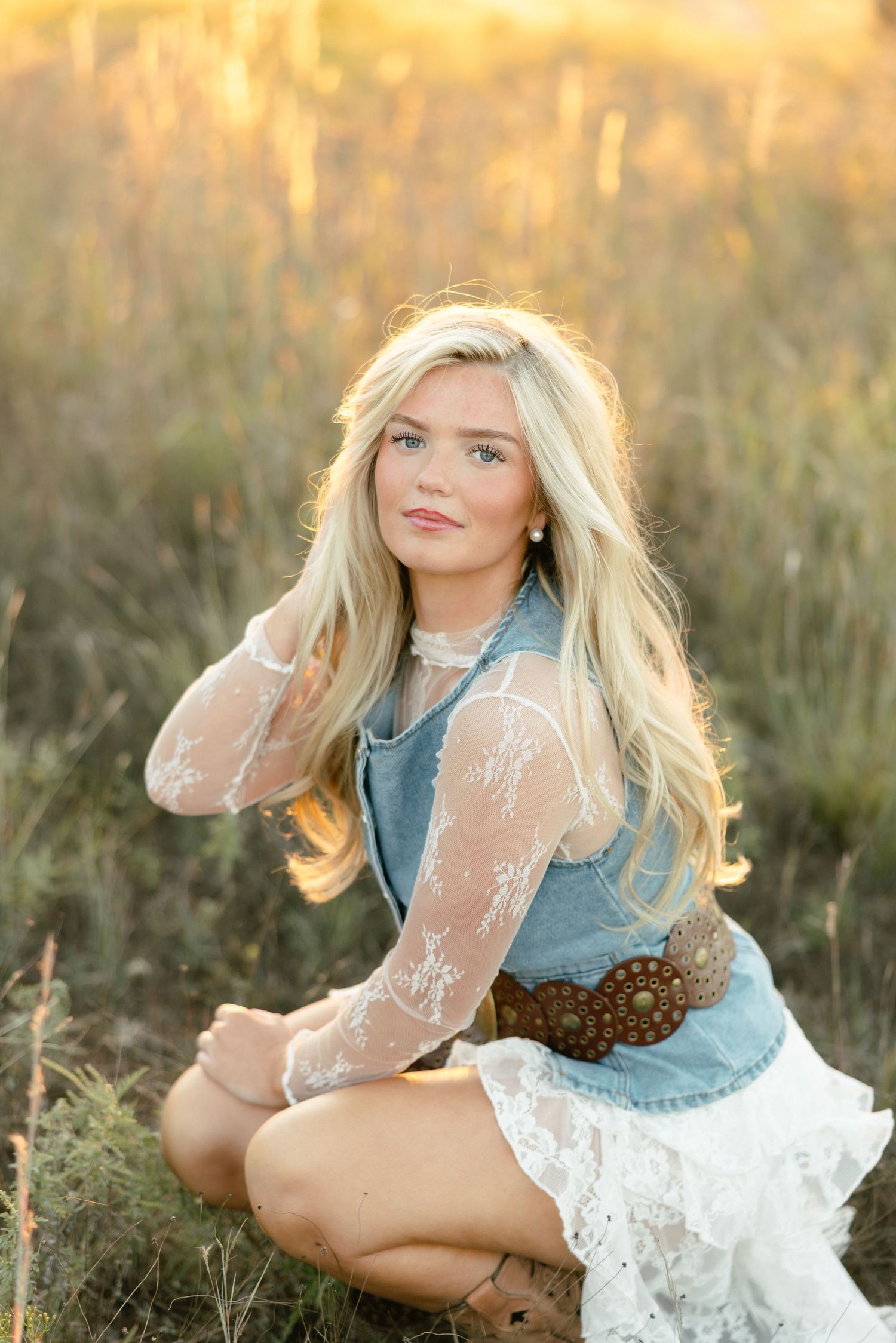 Smiling blonde girl in lace sleeves, denim vest, and brown belt sitting in golden grass field.