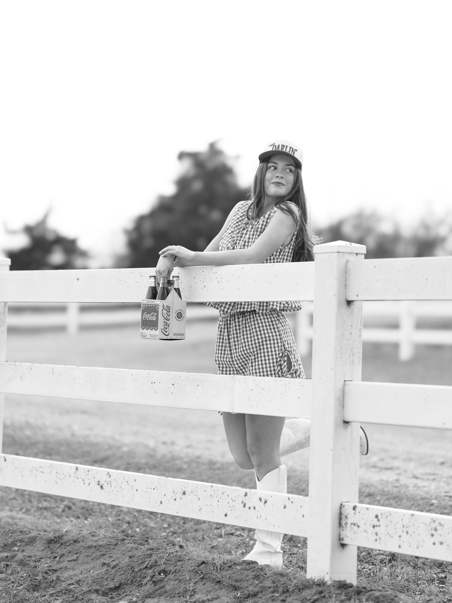 Young woman in checkered dress leaning on white fence holding Coca-Cola bottles, black and white.