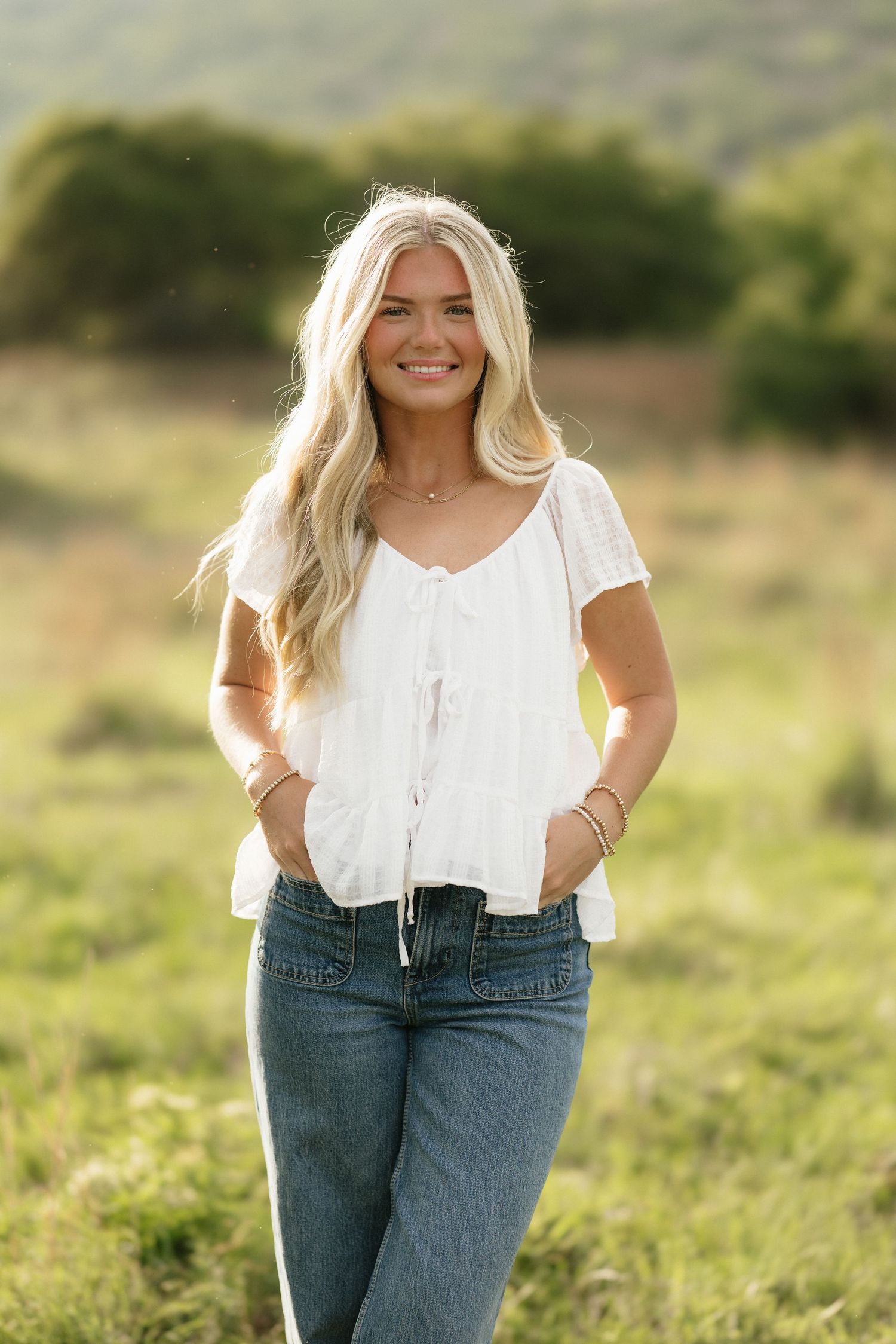 Smiling blonde woman in white top and jeans poses in a green field with mountains in the background.