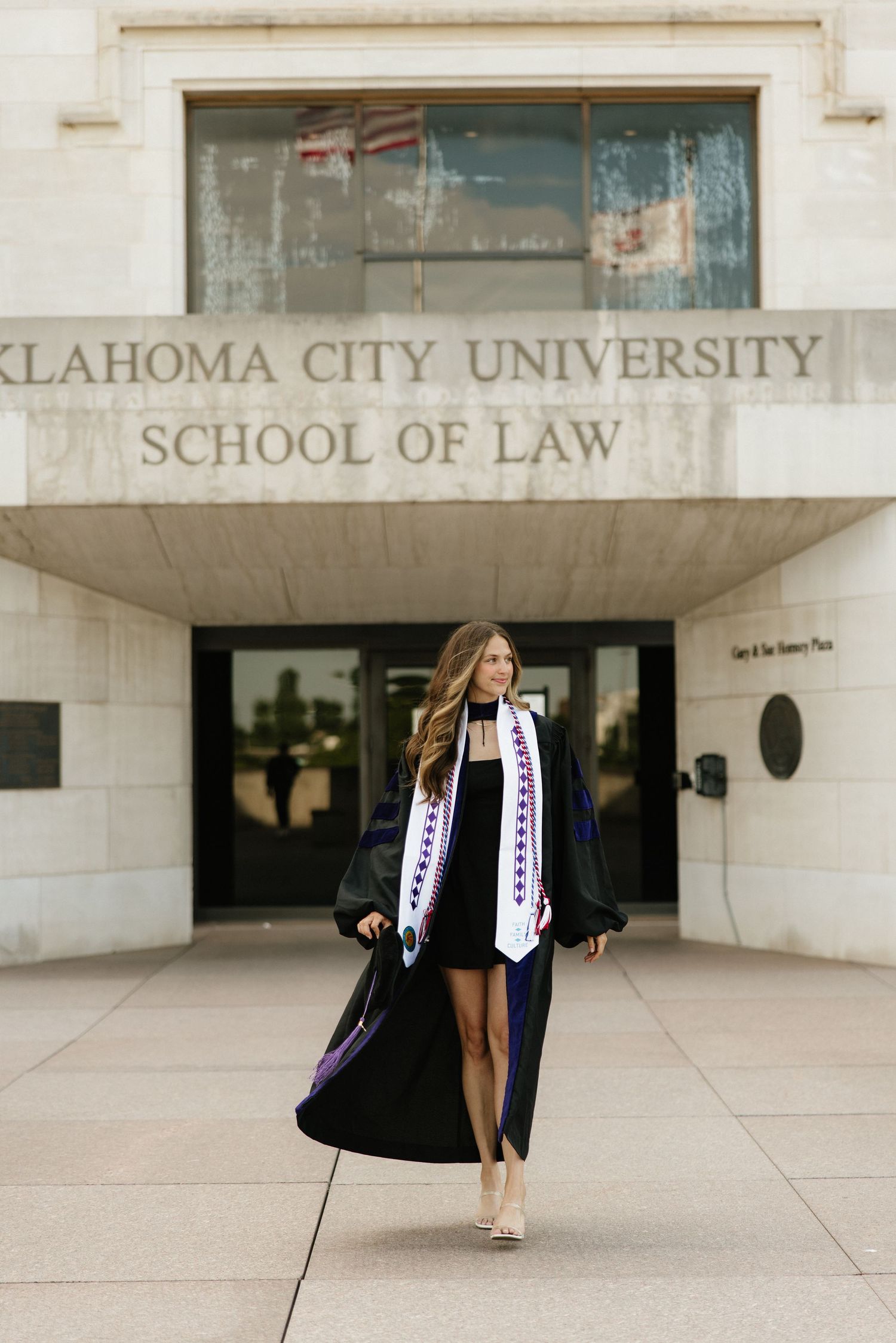 Woman in graduation cap and gown walking confidently outside Oklahoma City University School of Law.