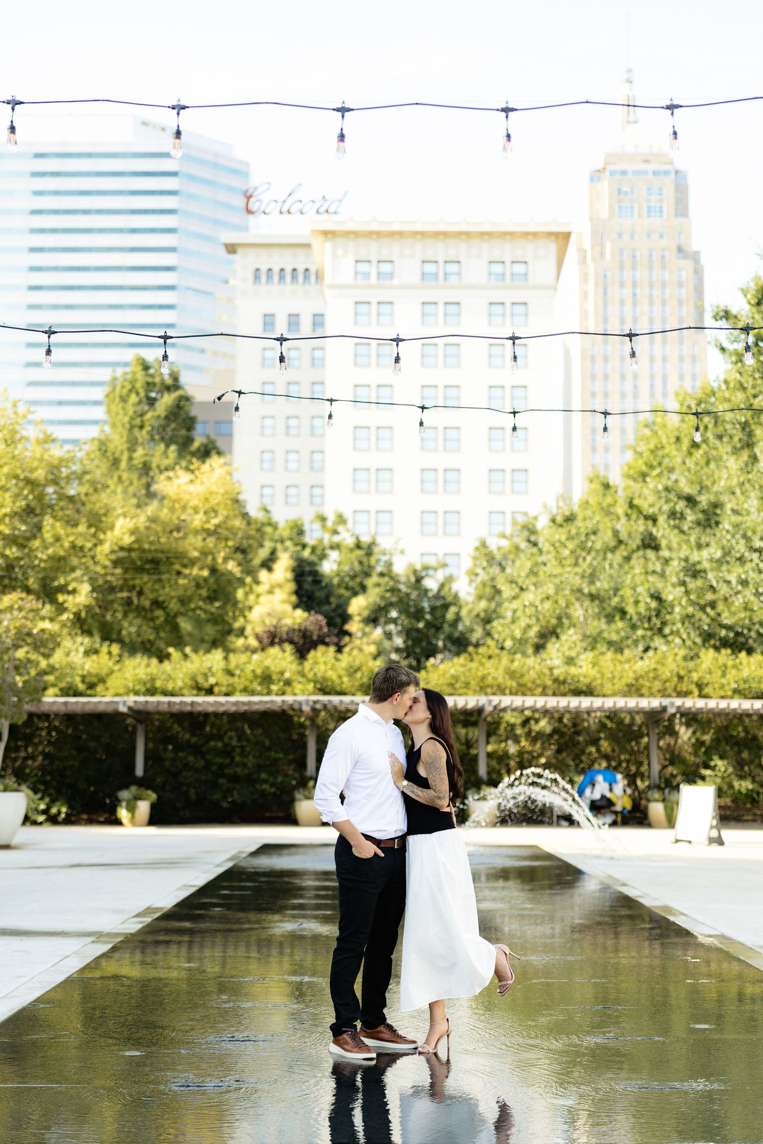 Couple sharing a romantic kiss by a fountain in an urban park with string lights and city skyline backdrop.