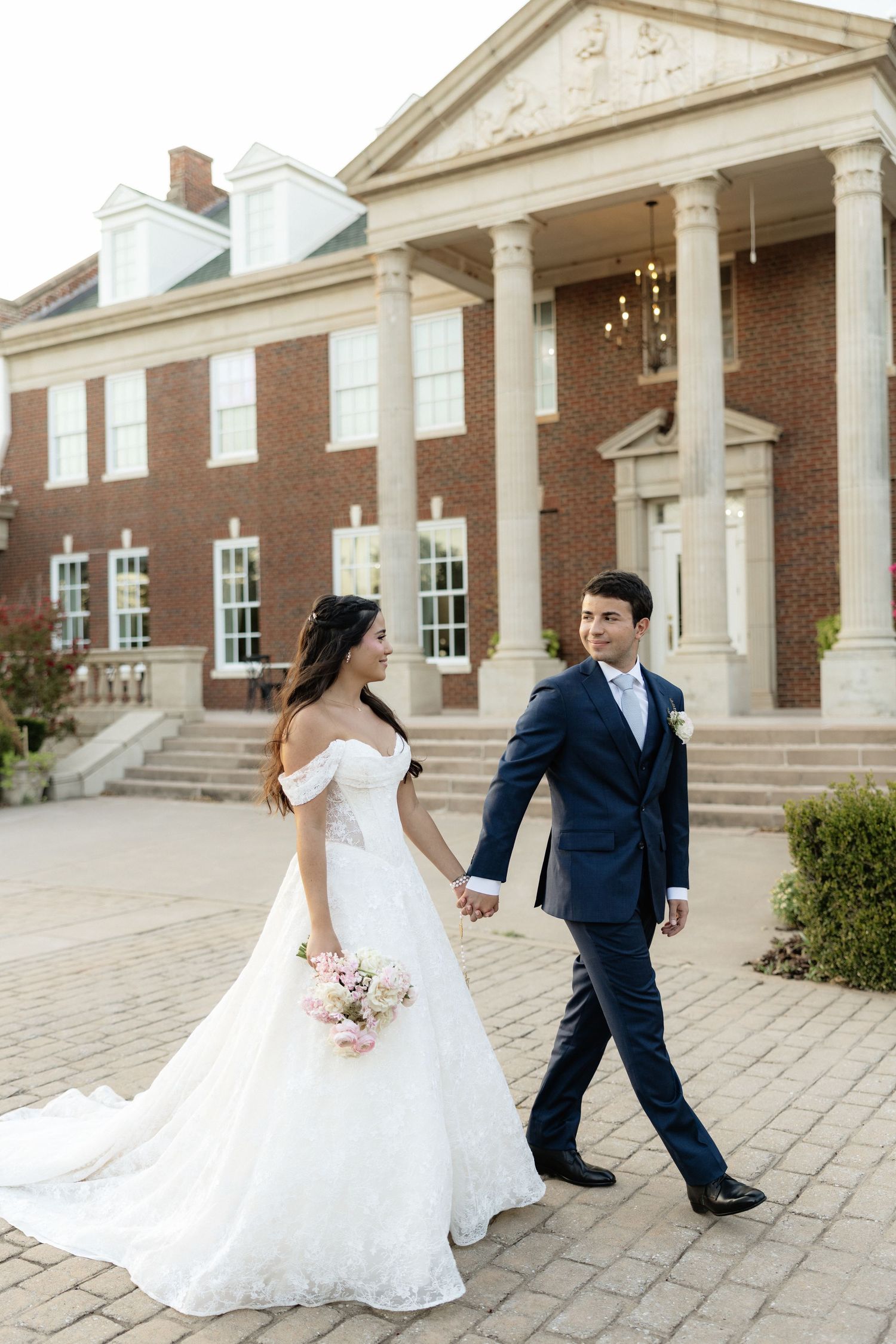 Bride and groom walk hand-in-hand before a grand brick colonial venue, she in a white lace gown, he in a navy suit.