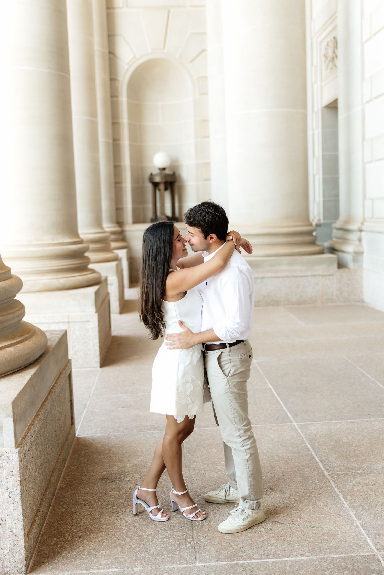 Couple embracing and almost kissing between elegant stone columns in a classic architectural setting.