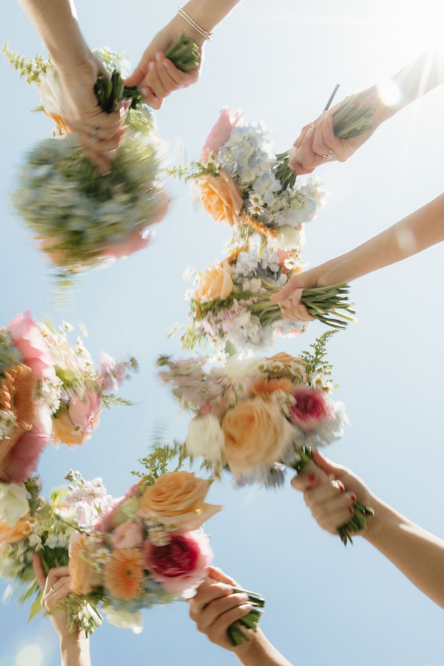 Bridesmaids holding colorful floral bouquets up toward blue sky photographed from below.