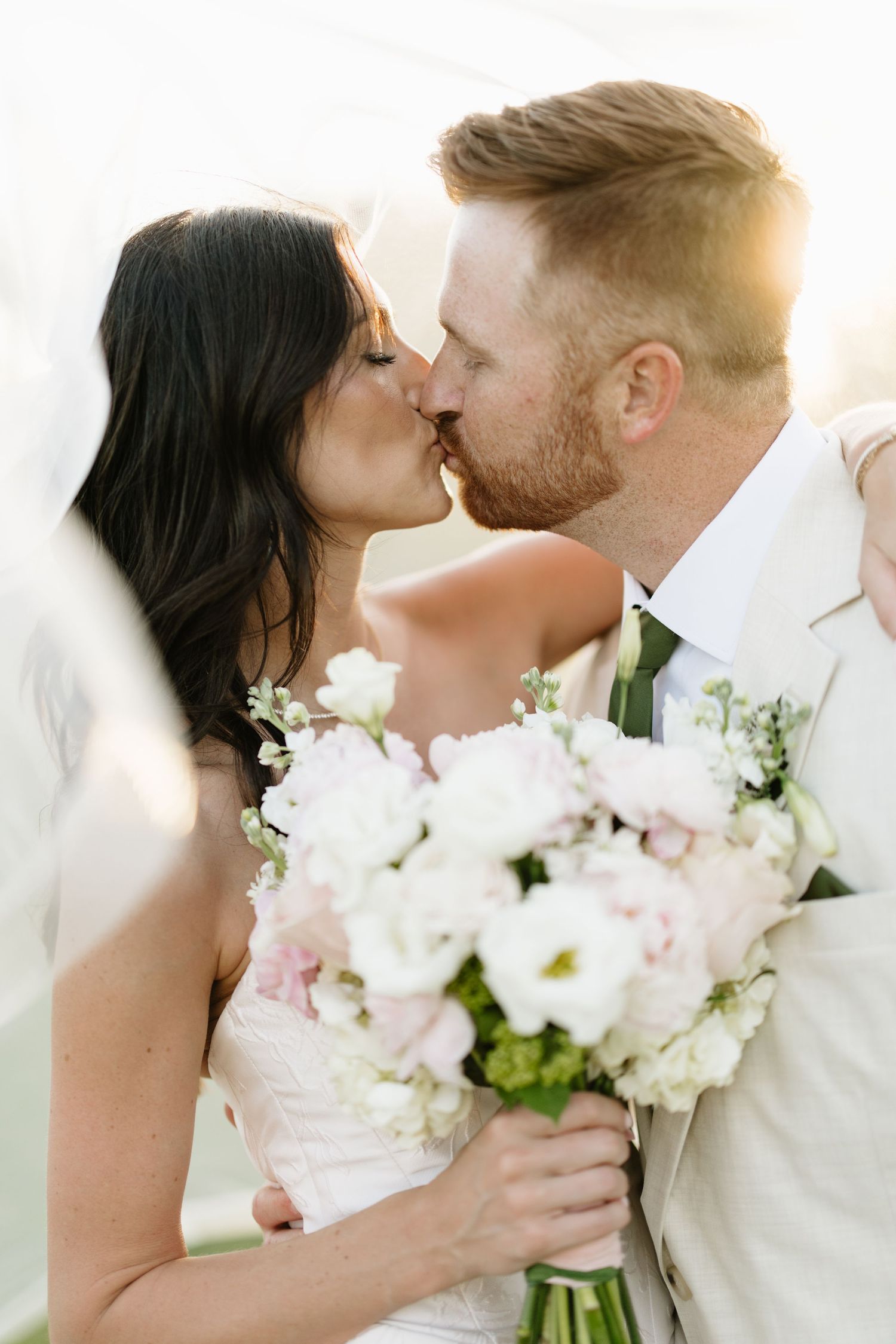 Bride and groom sharing a romantic kiss with white and pink bouquet in golden light.