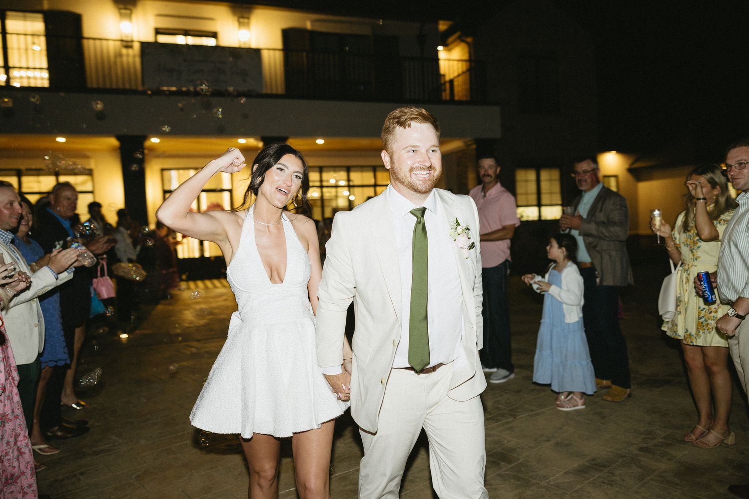 Newlyweds holding hands and dancing at outdoor evening wedding reception, groom in tan suit and green tie.