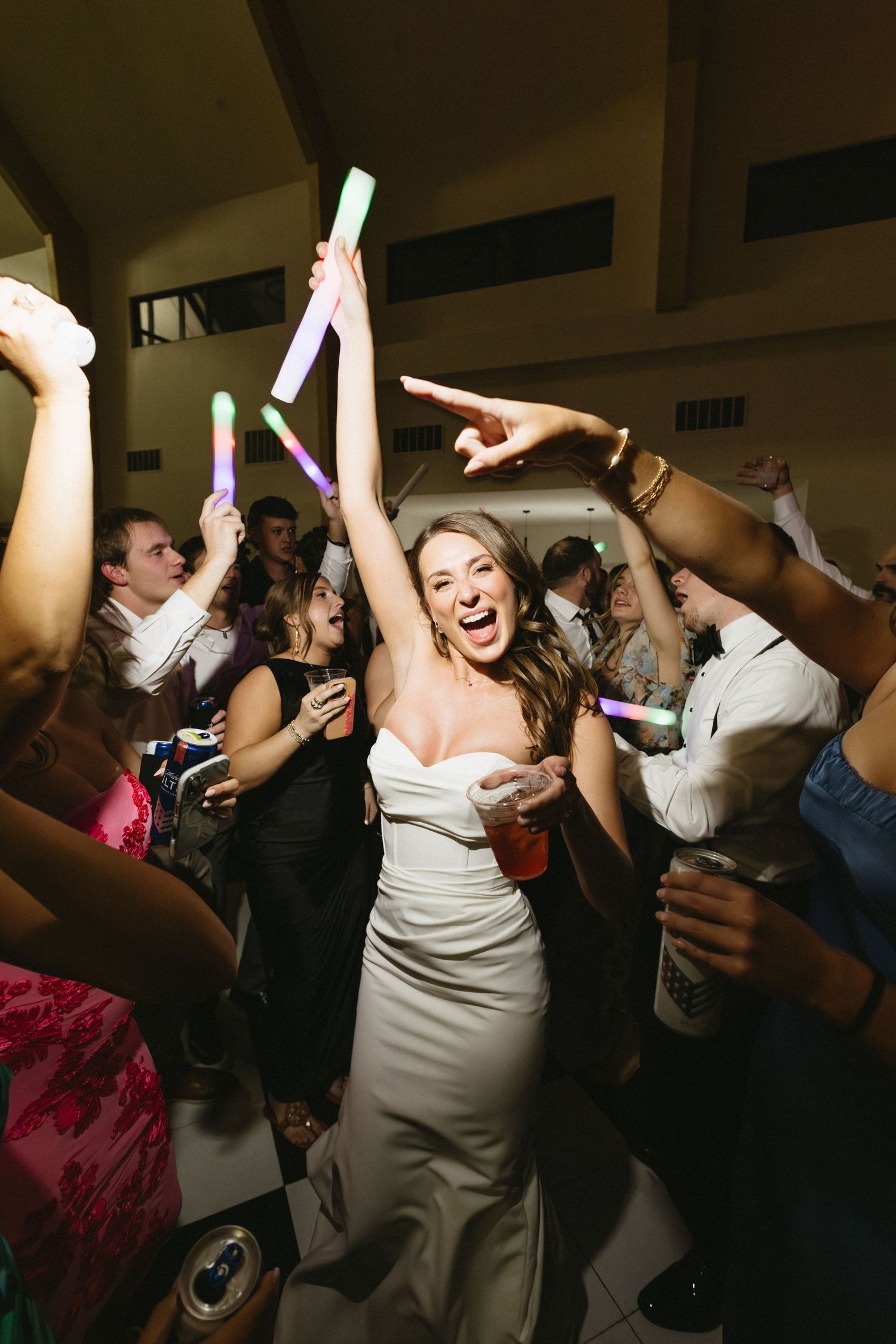 Excited bride in white gown celebrates on dance floor surrounded by guests with colorful glow sticks.