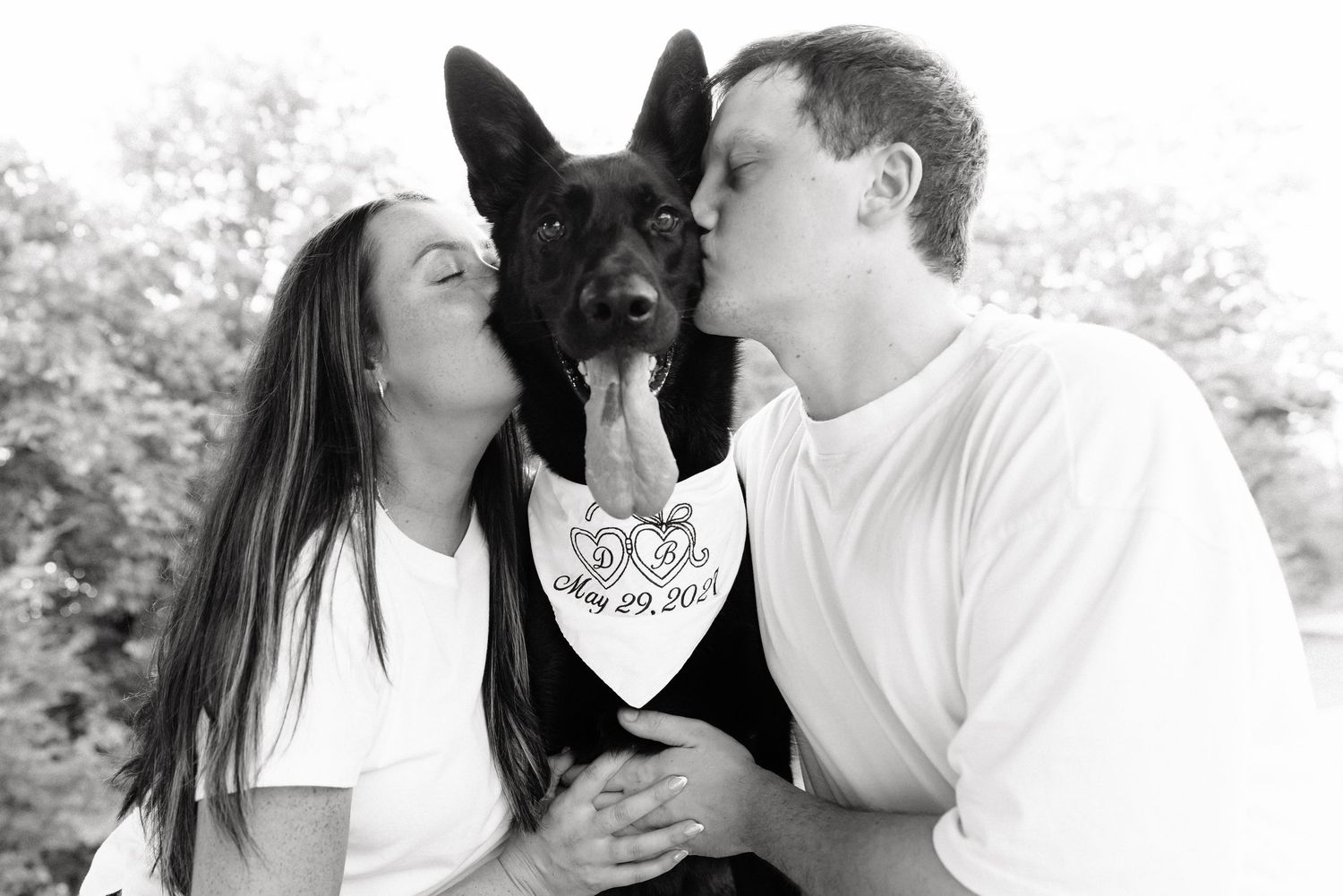 Couple kissing black German Shepherd dog wearing Save the Date bandana in black and white engagement photo.