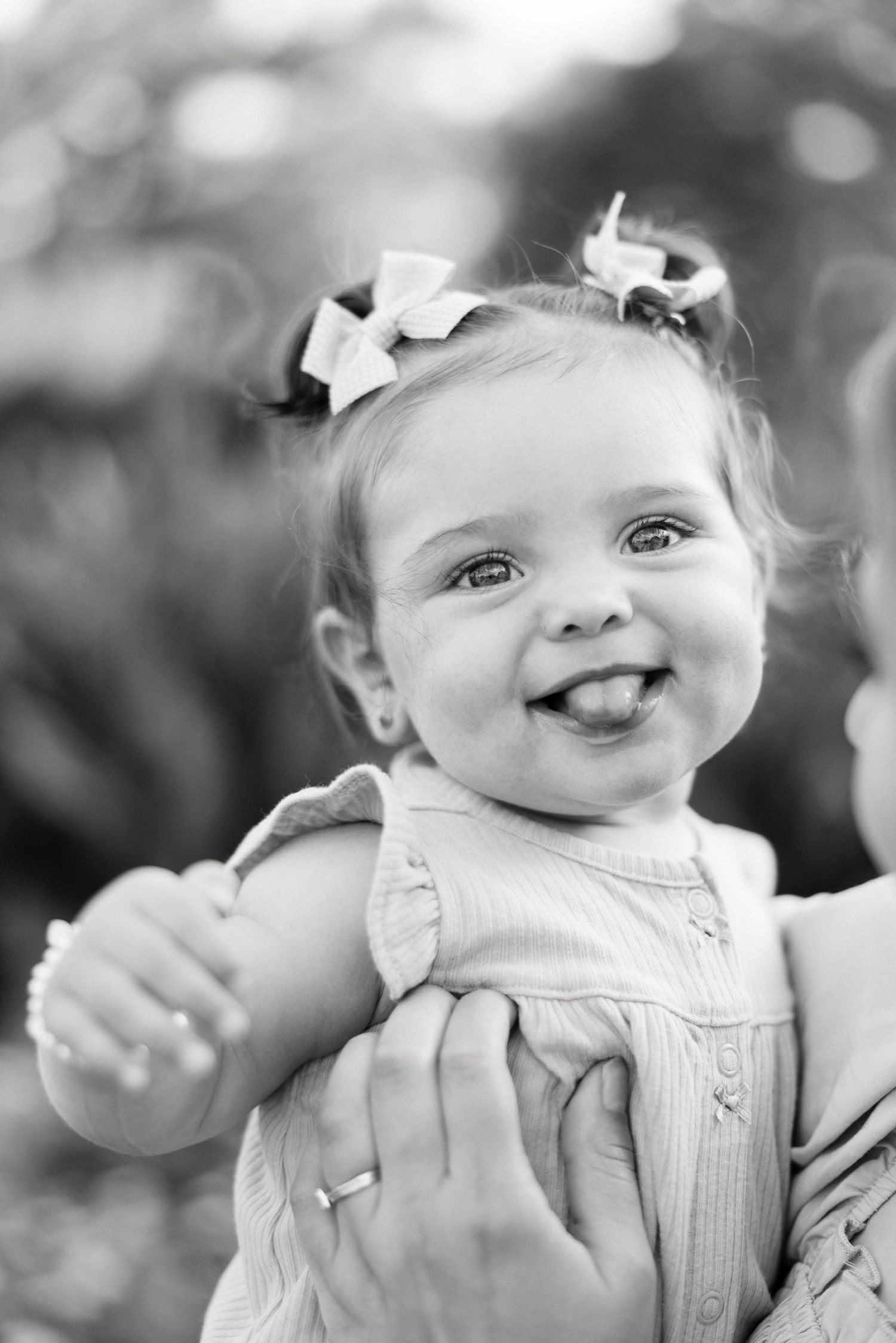 Black and white photo of a happy toddler girl with hair bows sticking out her tongue while being held by an adult.
