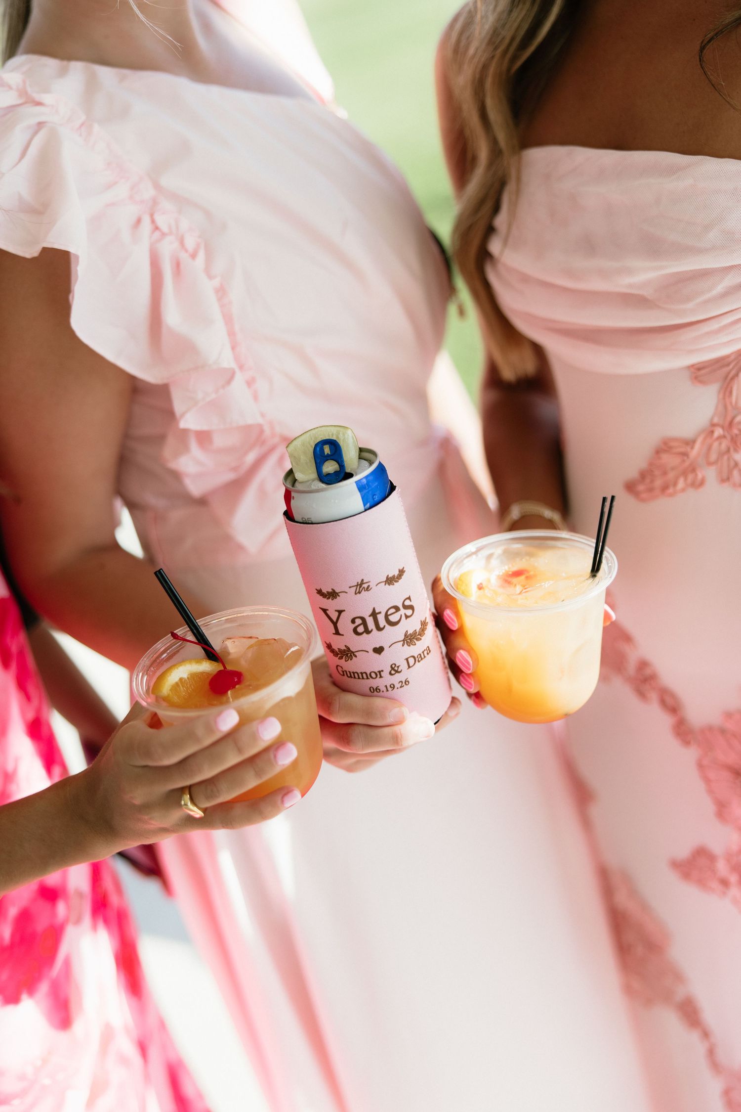 Bridesmaids in pink dresses holding cocktails and a personalized Yates wedding koozie at an outdoor celebration.