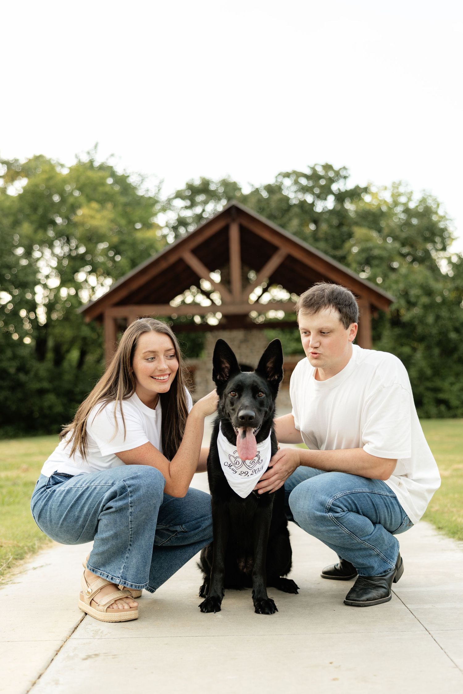 A couple in white tees and jeans crouch beside their black German Shepherd wearing a bandana in a park.