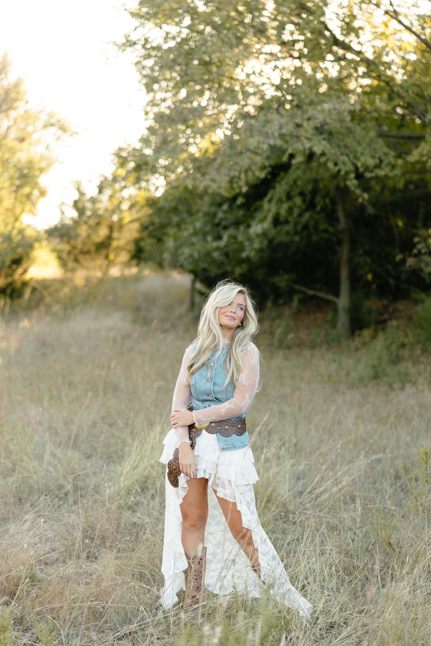 Blonde woman in boho denim vest, lace dress, and cowboy boots posing in golden meadow.