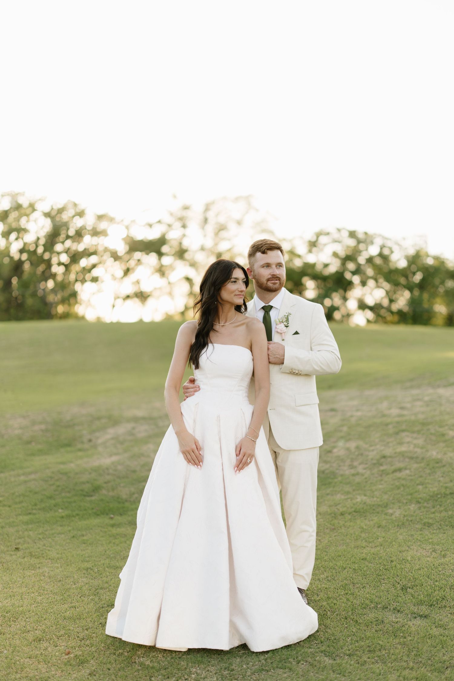 Bride in strapless white ball gown and groom in cream suit pose together on a golf course at sunset.