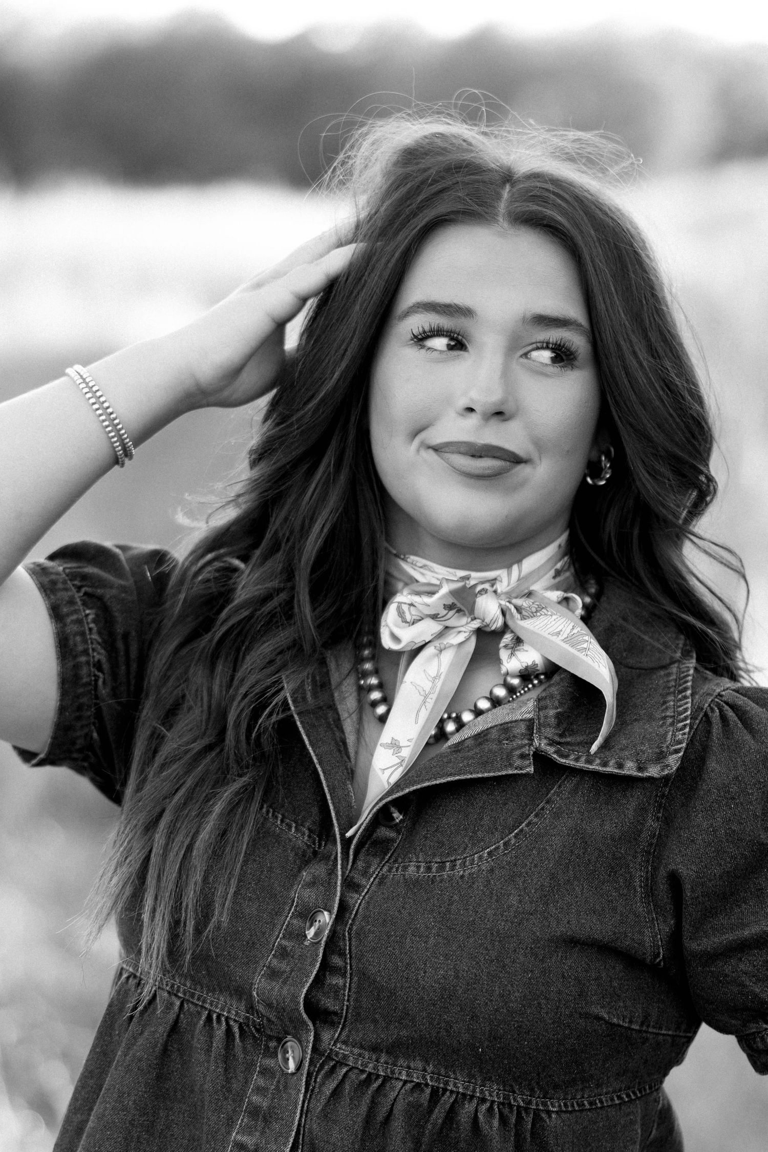 Black and white portrait of a young woman in denim dress with neck scarf touching her hair.