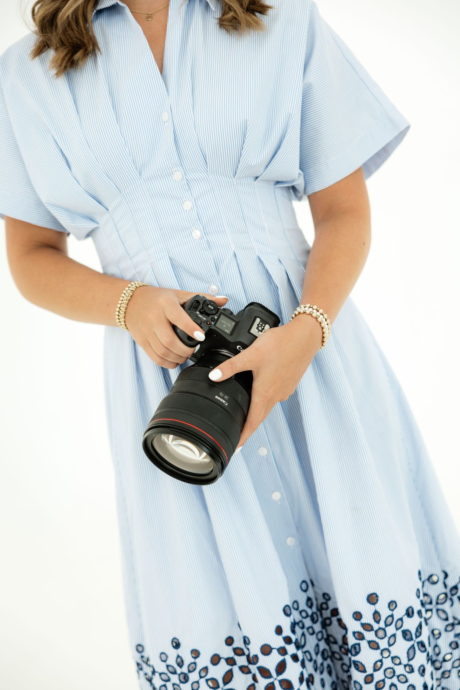 Woman in light blue floral dress holding a Canon DSLR camera with pearl bracelets on white background.