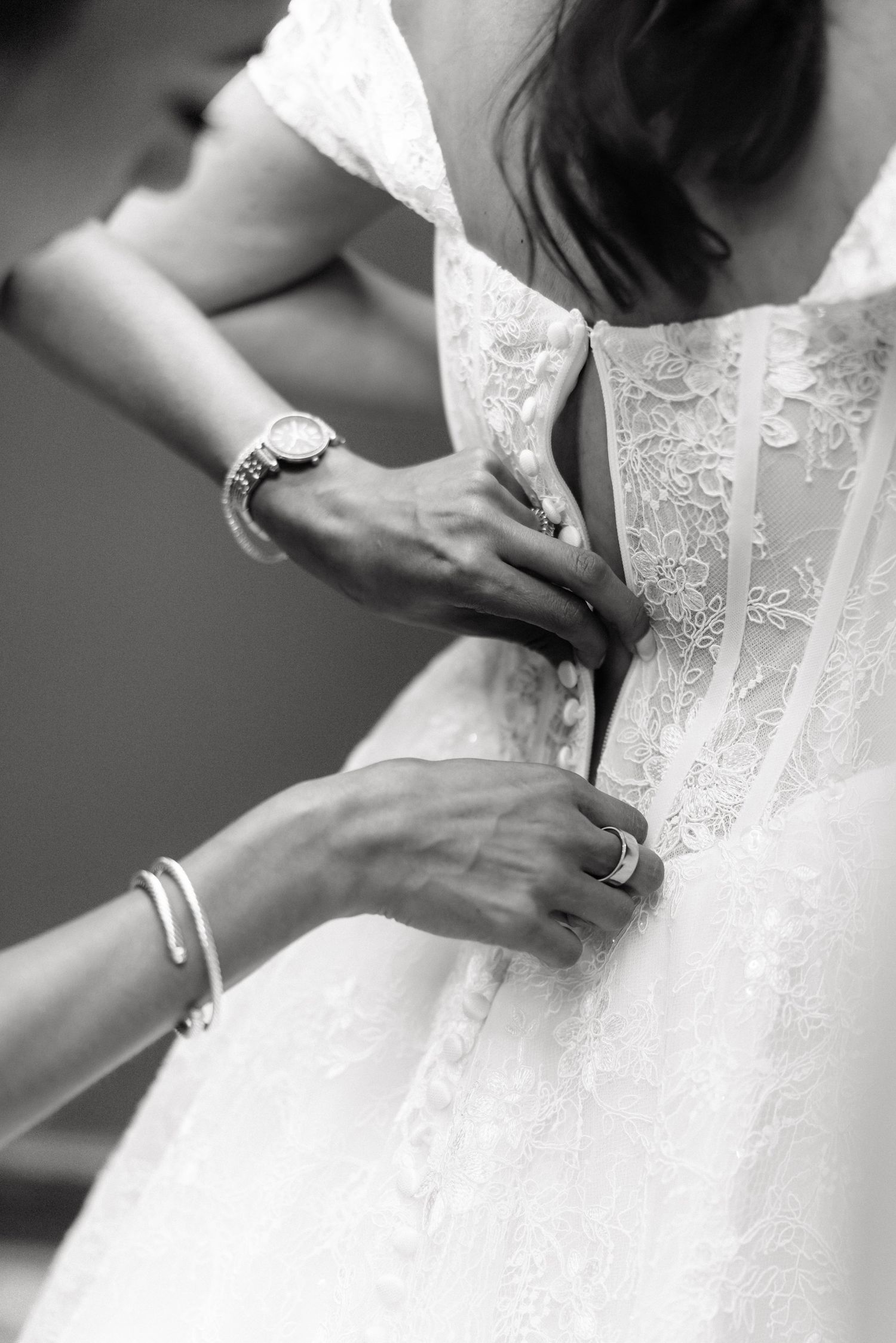 Black and white photo of hands buttoning the back of a lace wedding dress, with bridesmaid wearing watch and bracelet.
