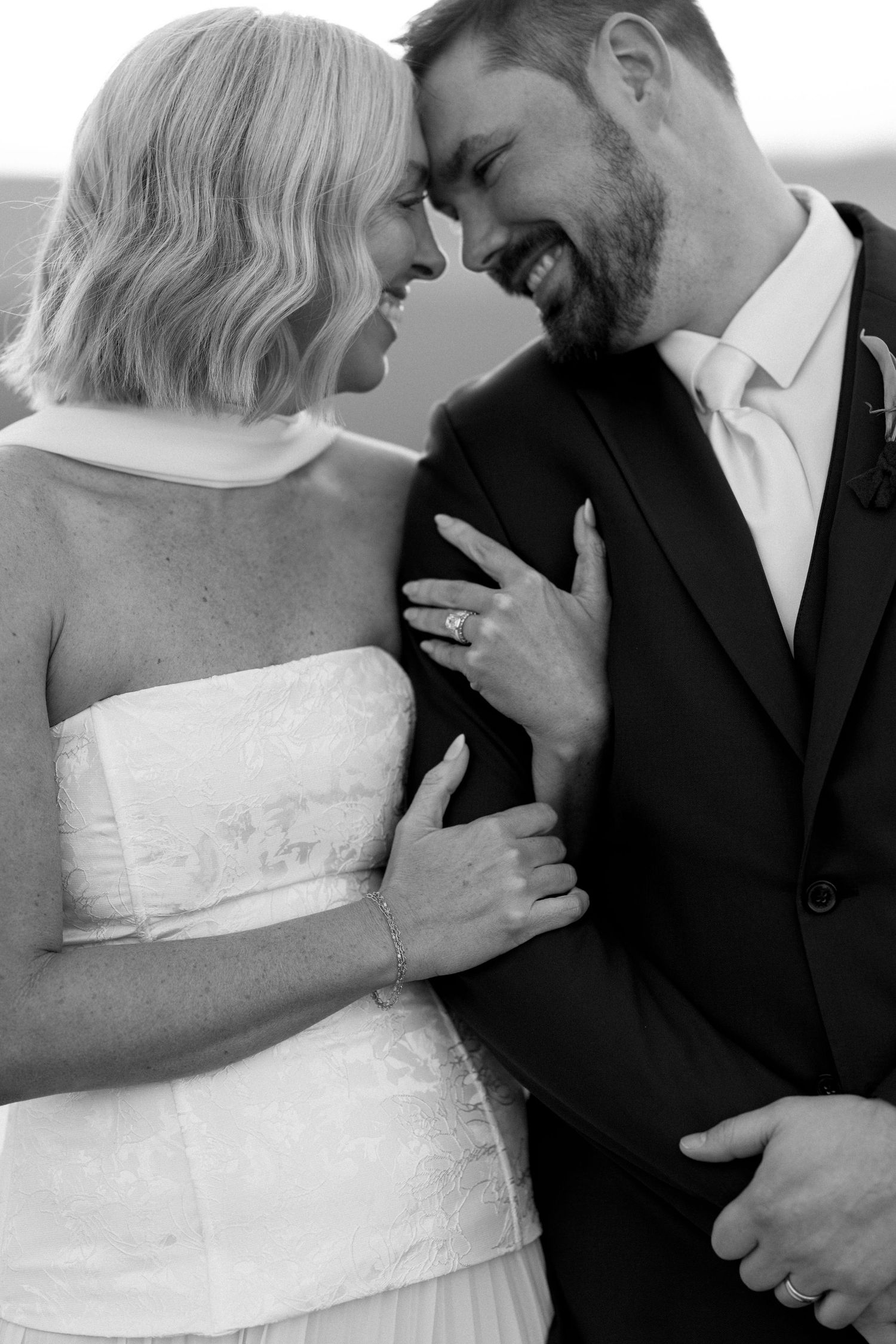 Black and white portrait of smiling newlyweds touching foreheads, bride showing ring.