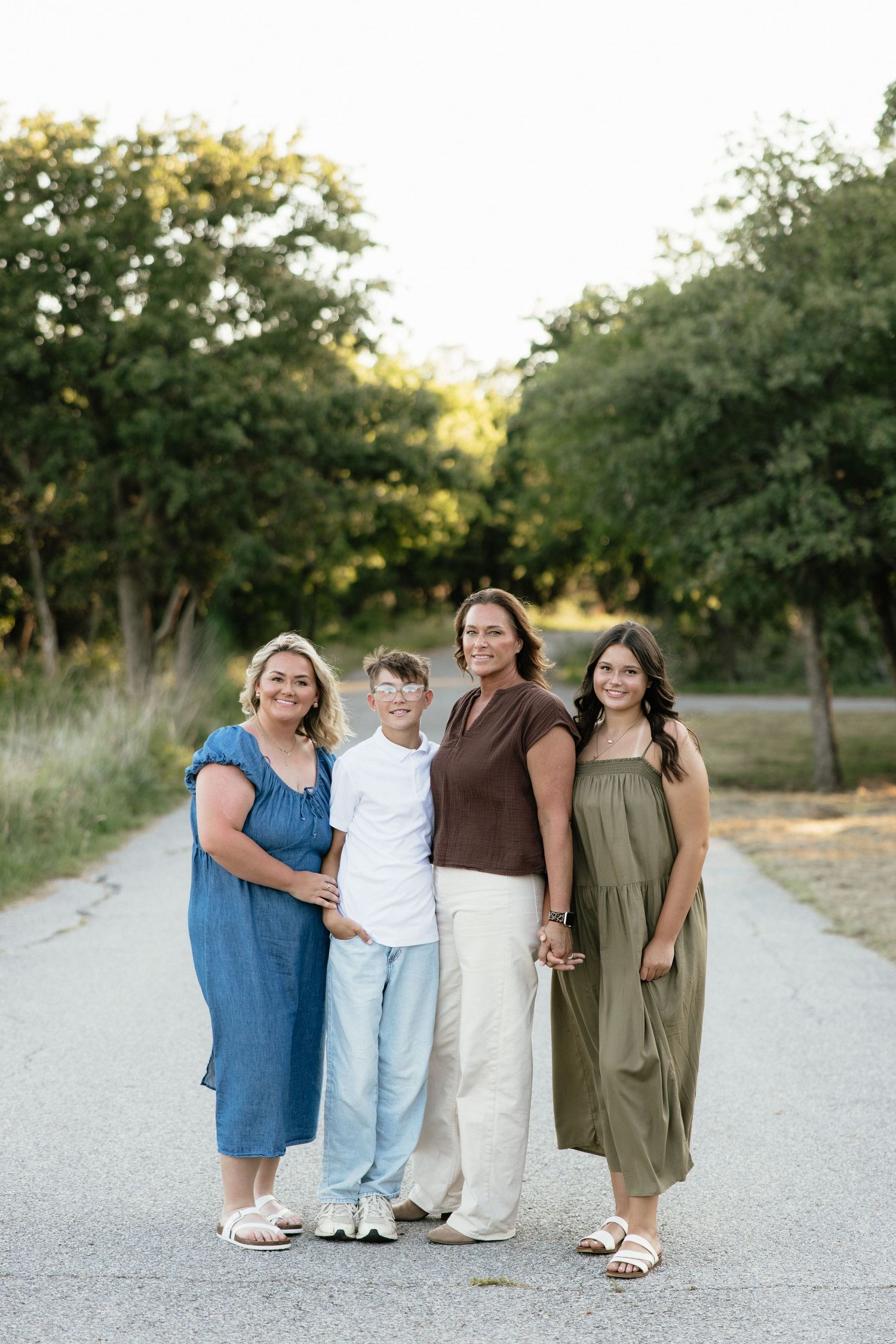 Family of four posing on a tree-lined path during golden hour, wearing coordinated outfits in blue, white, brown, and olive green.