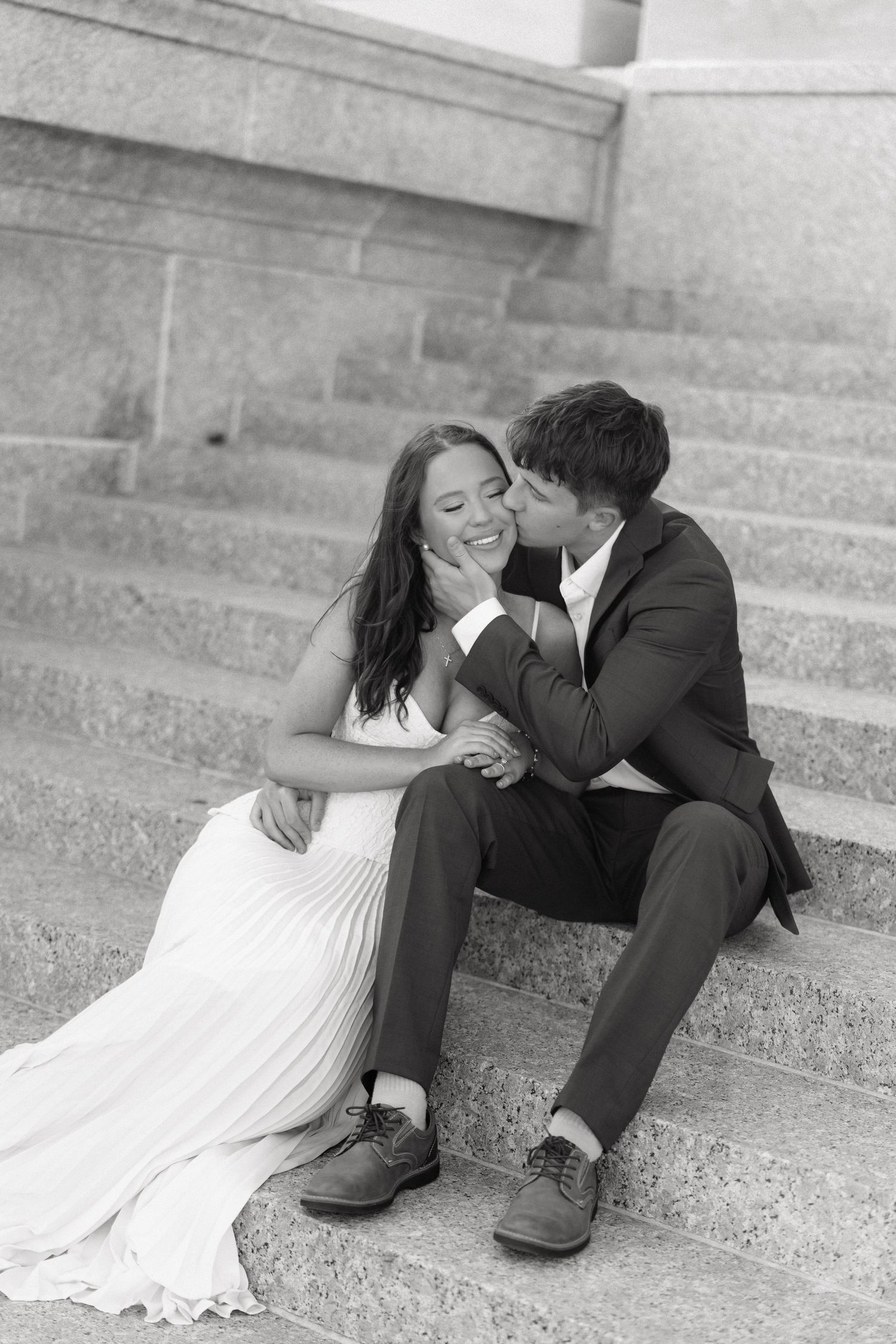 Black and white photo of a groom kissing his bride on the cheek as they sit together on stone steps.
