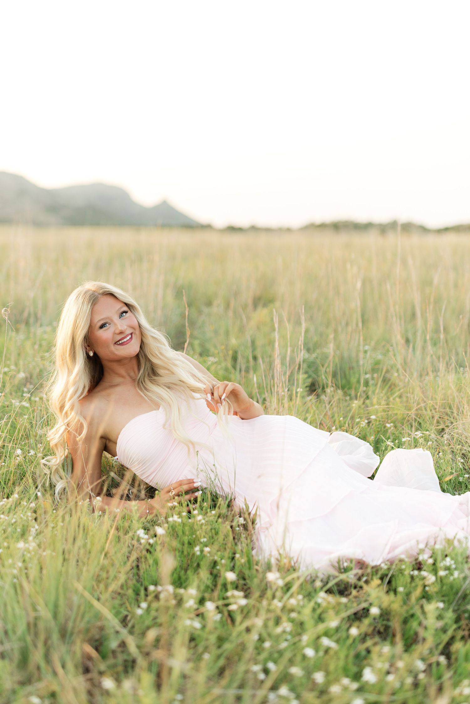 Blonde woman in white dress lying in green field with mountains, smiling upward in soft natural light.