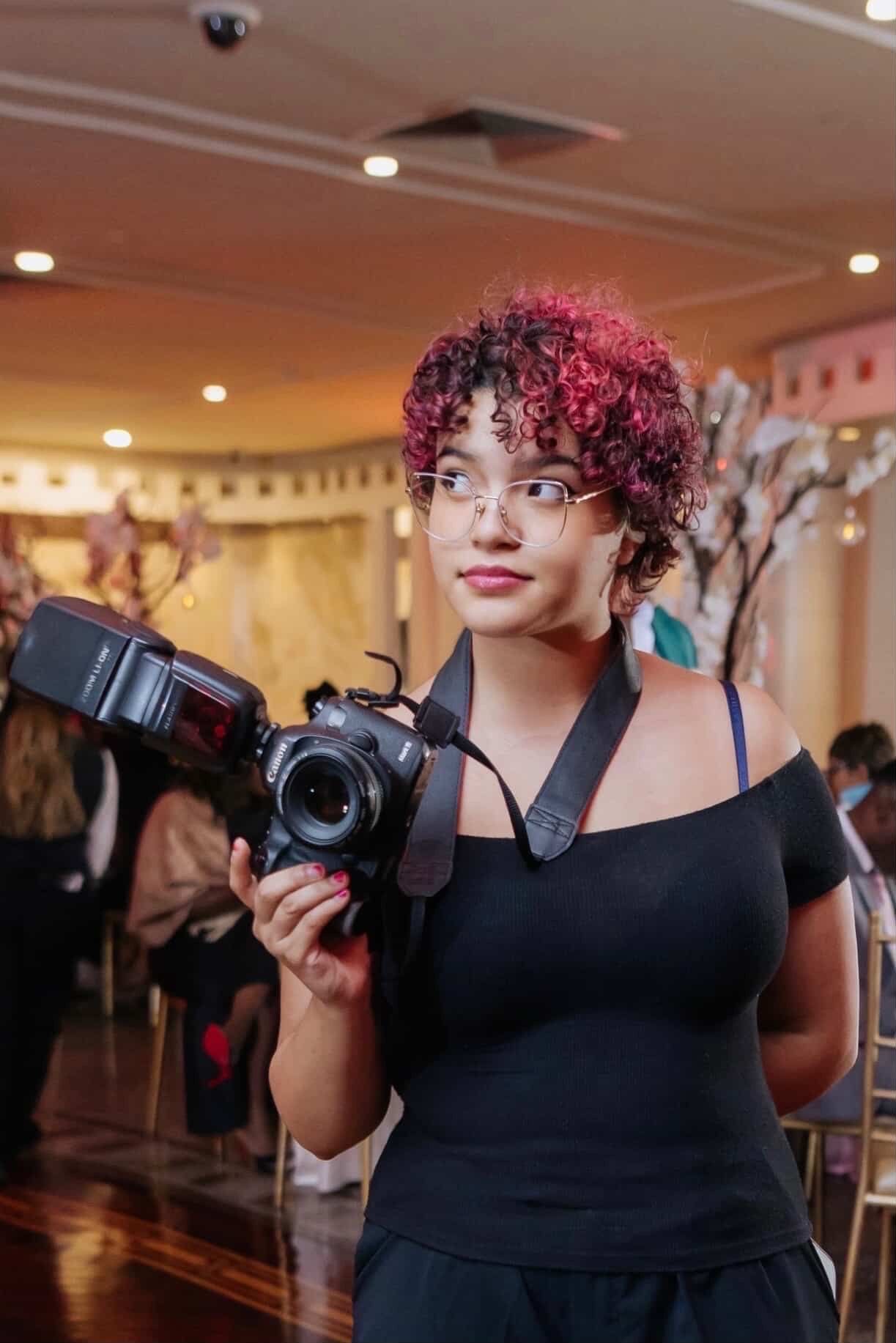 Professional photographer with pink curly hair wearing black dress holds camera at indoor event venue.