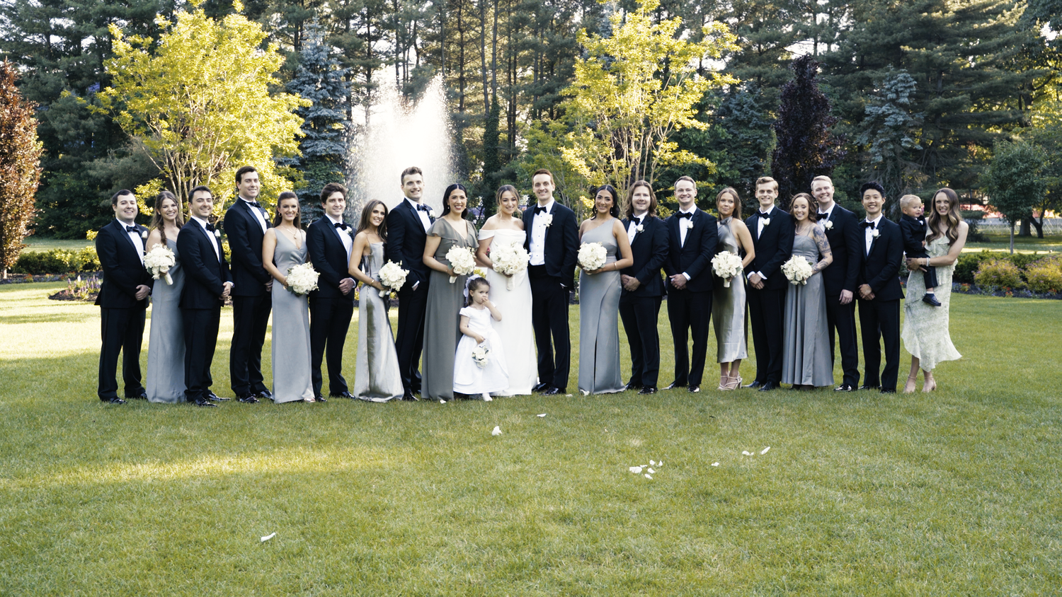 Wedding party poses in a line on grass with trees and sunlight in the background.