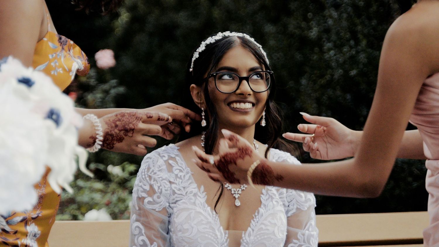 A joyful bride in a lace dress and glasses smiles while being helped with final touches.